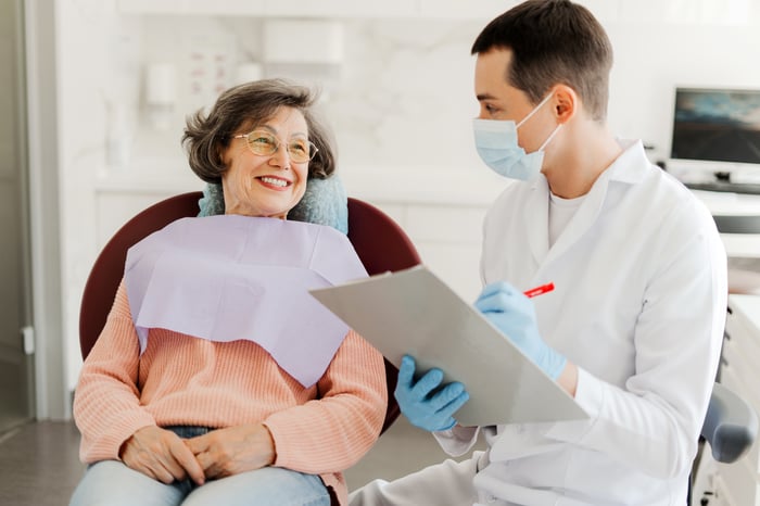 An older woman smiling in a dental chair while her dentist reviews a treatment plan on a clipboard.