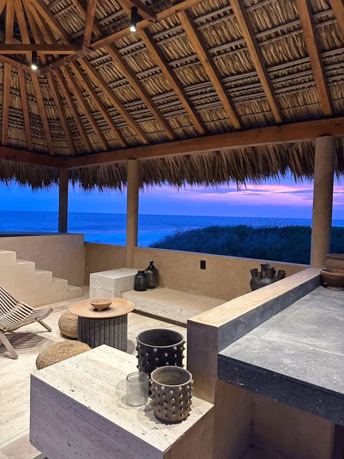 Outdoor seating area under a thatched roof with a view of the ocean at sunset, featuring stone furniture and decorative pottery.