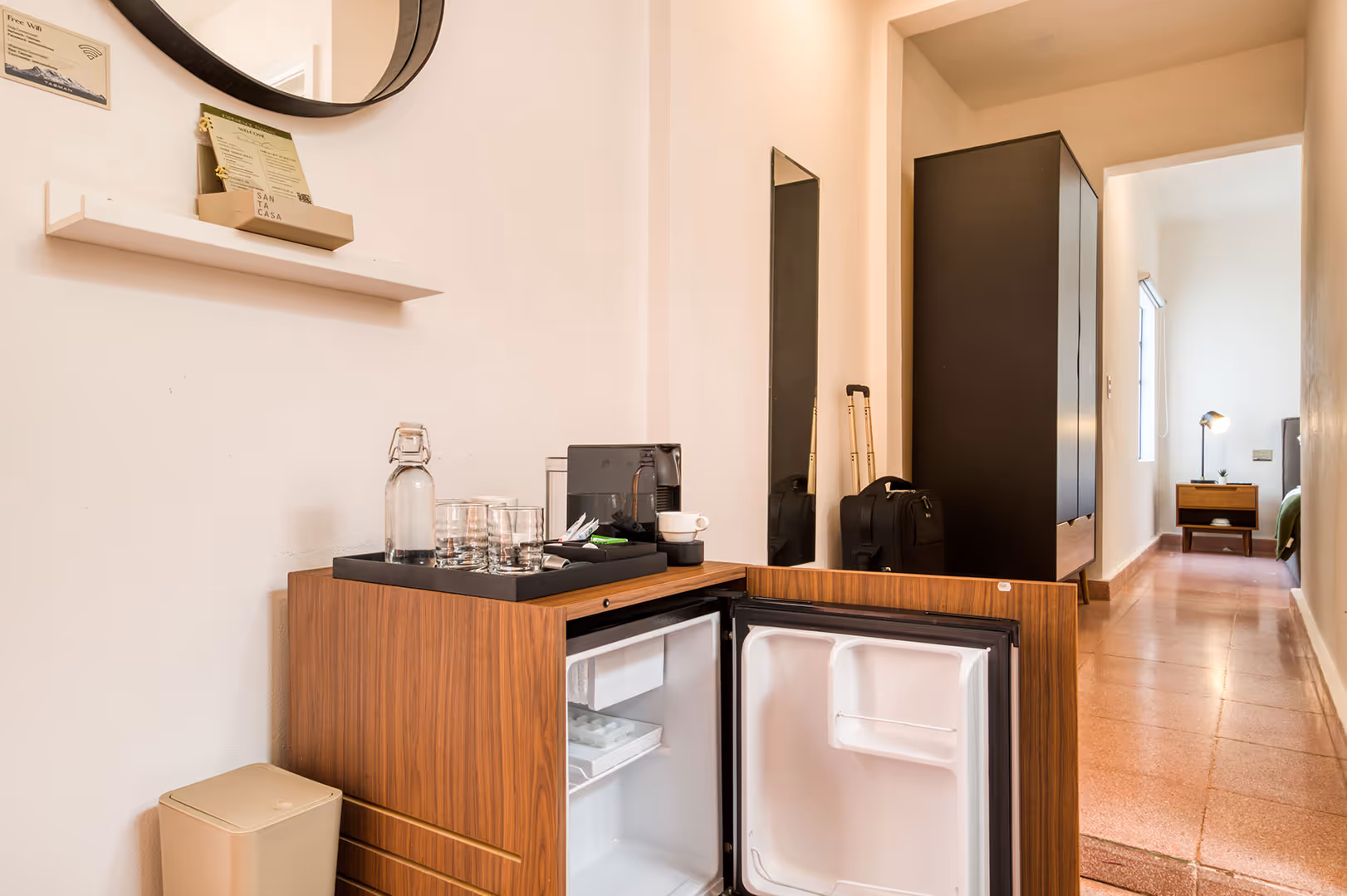 Hotel room corner with open mini fridge in wooden cabinet, water bottle, glasses, coffee machine on top, and hallway leading to a bedroom with bedside table and lamp.