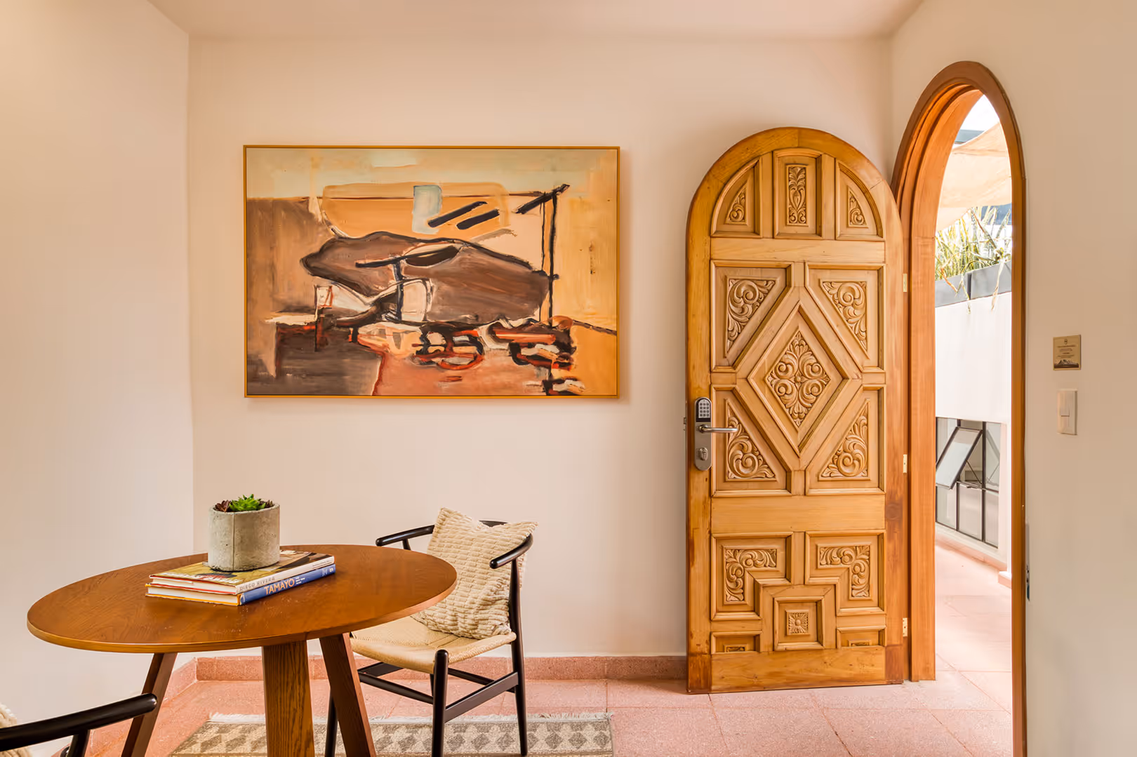 Bright room with a carved wooden arched door, abstract painting on white wall, round wooden table with books and succulent, and a cushioned chair.