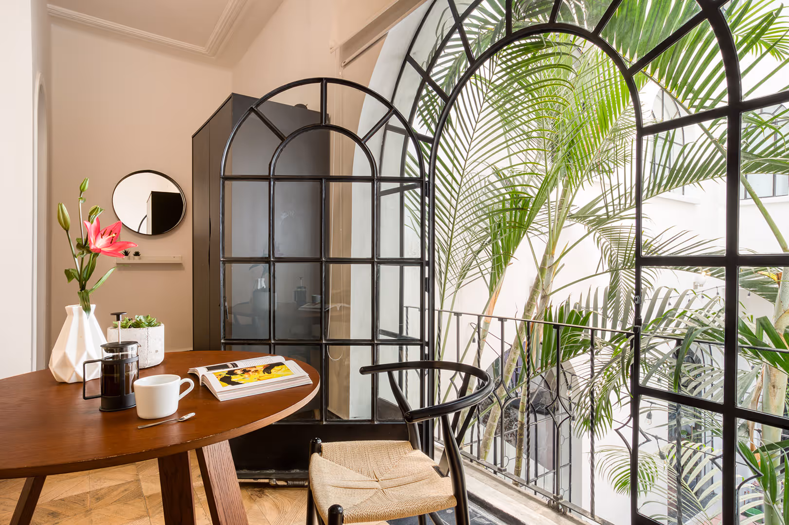 Cozy interior space with a round wooden table set with a coffee press, cup, open book, and flowers, next to an open black-framed arched door revealing green palm plants outside.