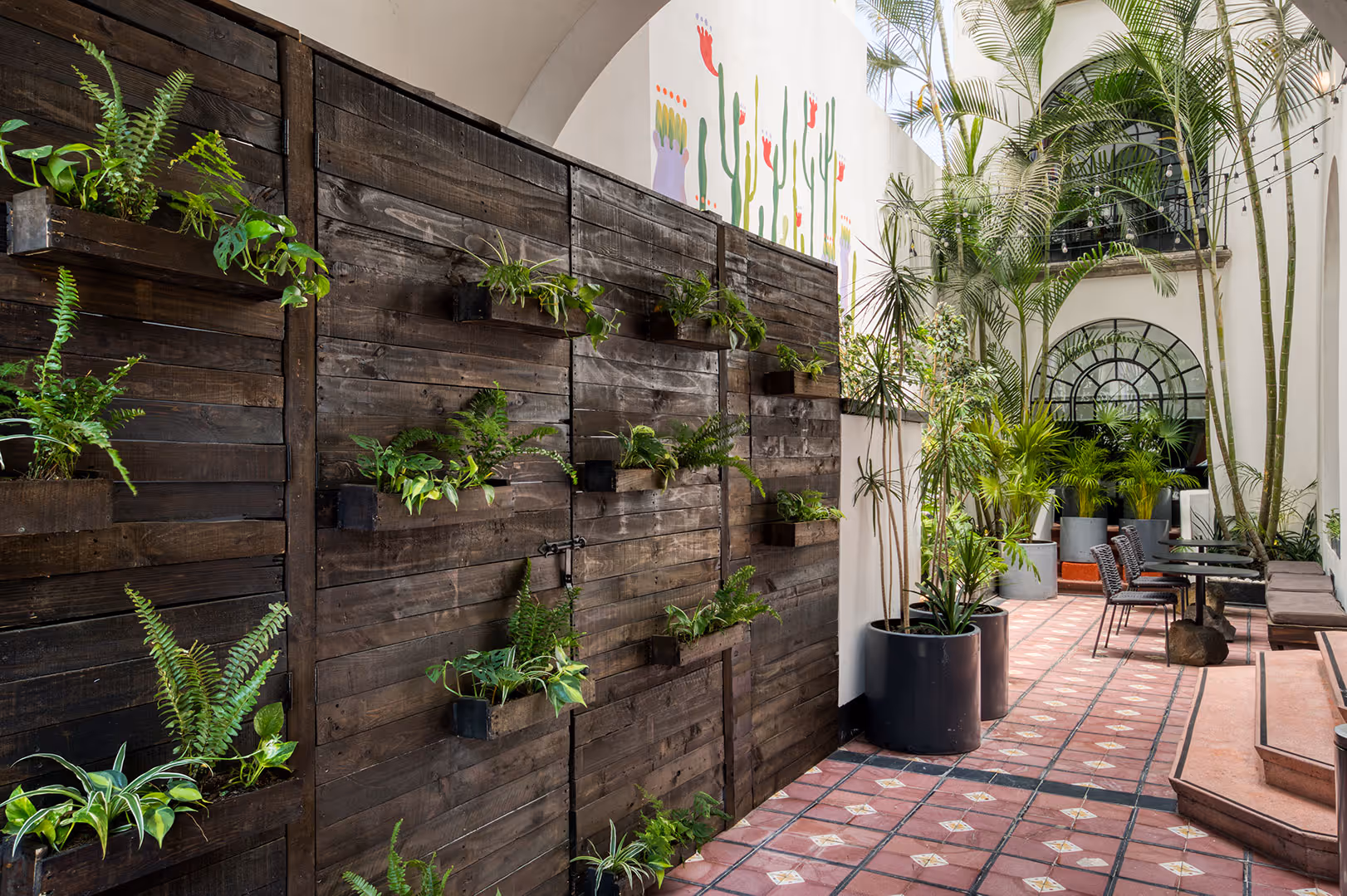 Indoor courtyard with tiled floor, tall potted plants, a wooden wall-mounted planter with green foliage, and a seating area with chairs and tables.