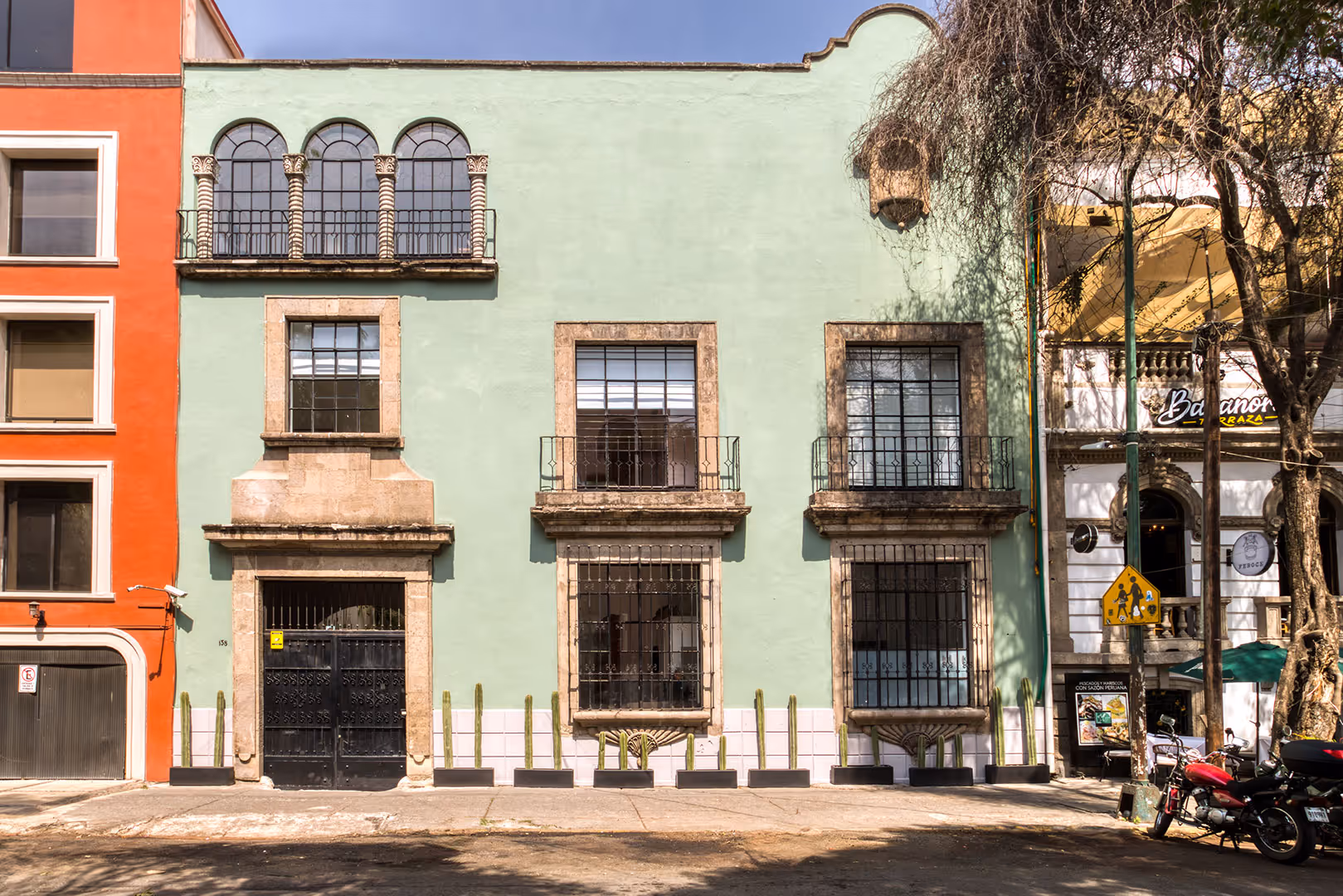 Facade of a light green building with arched windows and decorative stone frames, flanked by an orange building and a white building with a terrace.