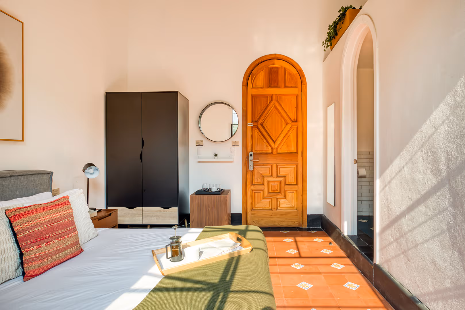 Bright bedroom with a wooden arched door, black wardrobe, bed with colorful pillows, and sunlight casting shadows on tiled floor.