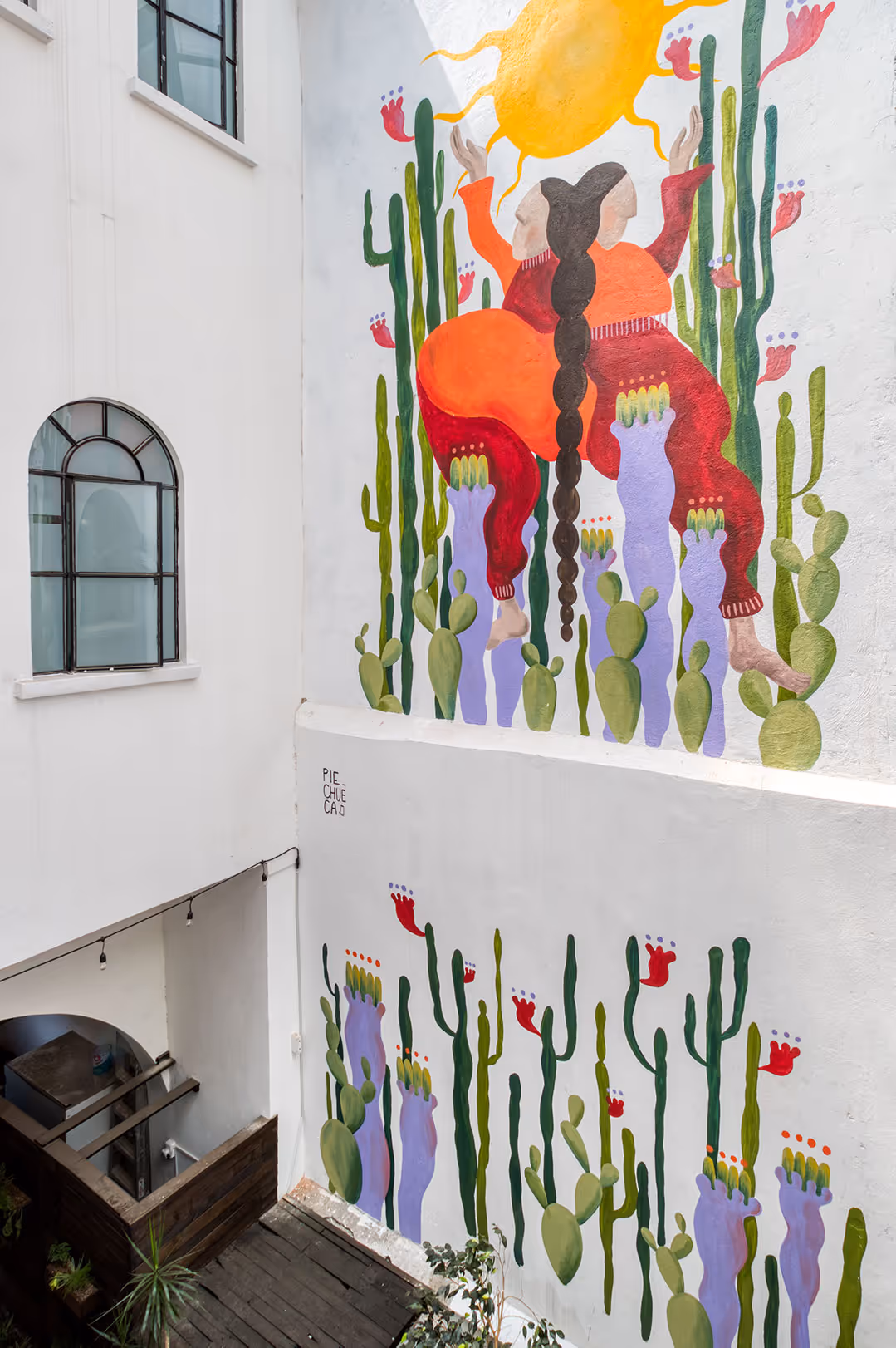 Colorful mural on white wall depicting a person with long braided hair and red clothing among cacti and plants.