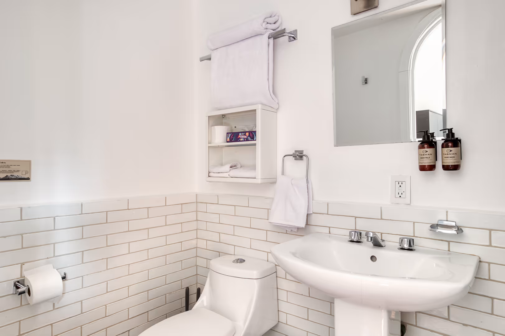 Clean bathroom with white tiled walls, a toilet, pedestal sink, mirror, mounted soap and body milk dispensers, and neatly folded white towels on a rack and shelf.