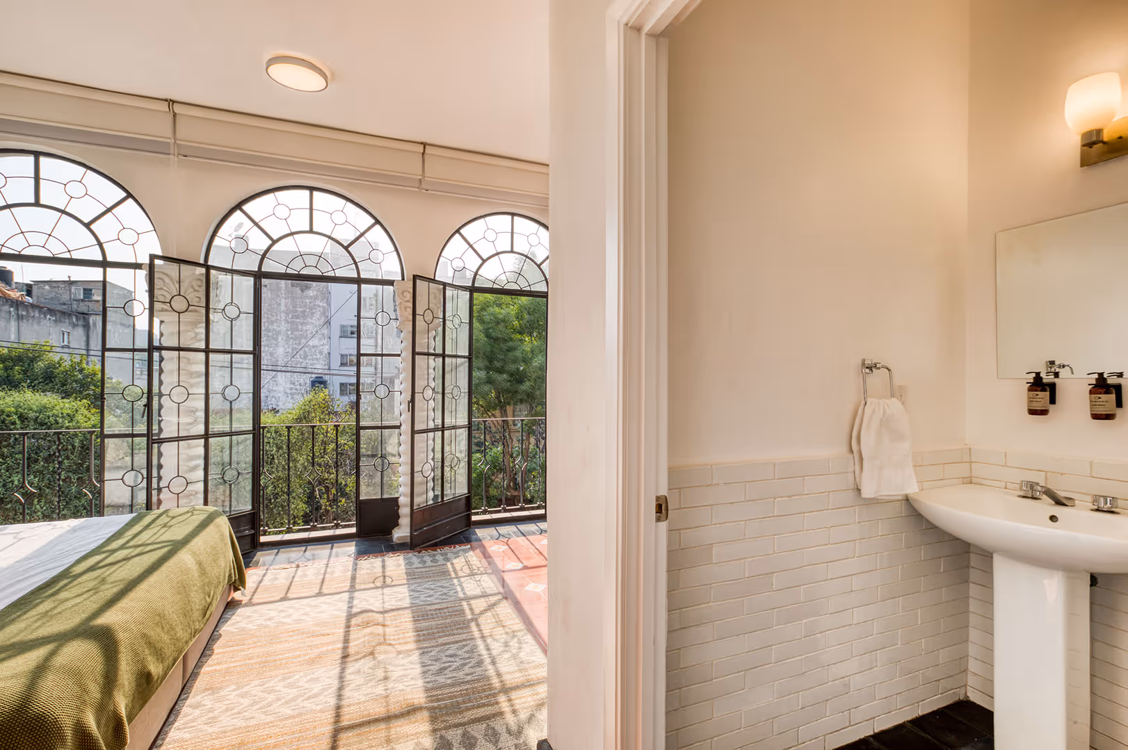 Sunlit bedroom with a green bedspread and arched glass doors opening to a balcony, adjacent to a bathroom with a pedestal sink and towel.