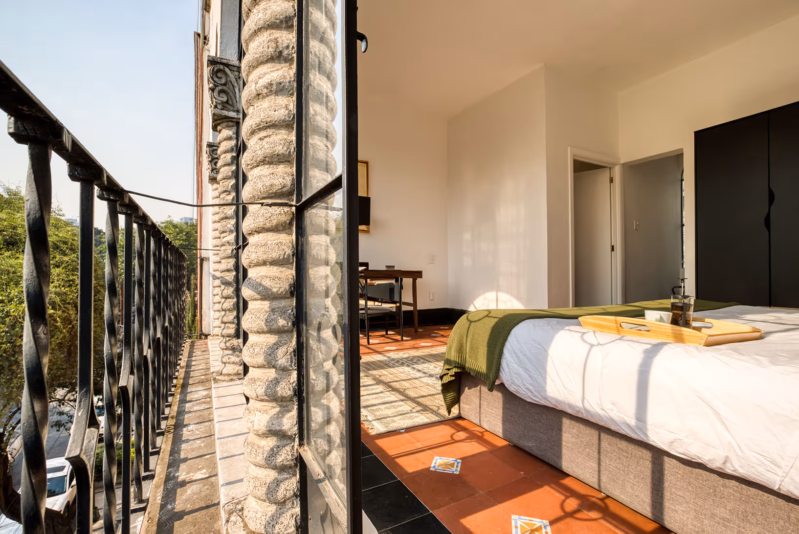 Sunny bedroom interior with a bed covered by a white duvet and green throw, a wooden tray on the bed, black wardrobe, and a small balcony with wrought iron railing.