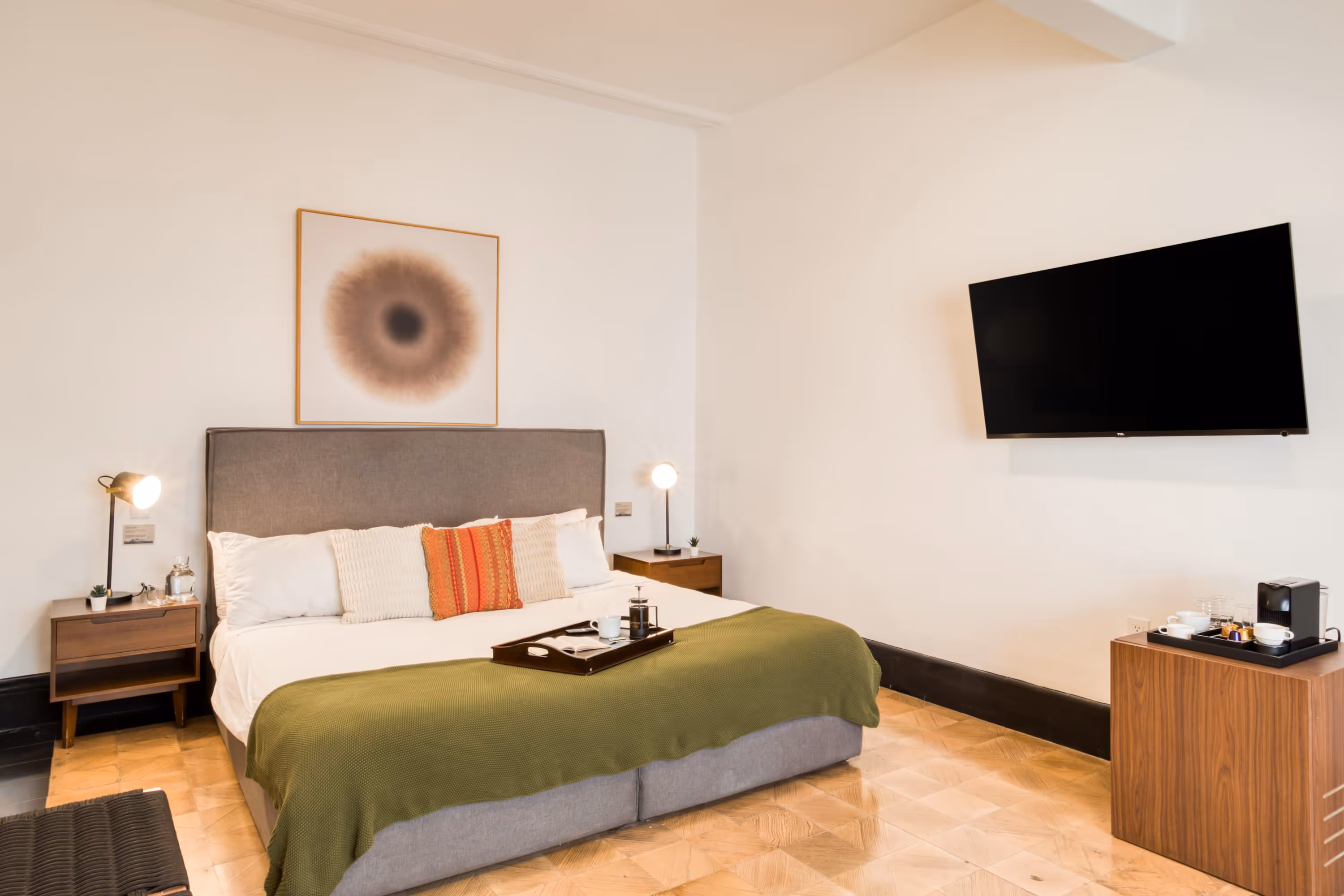 Modern bedroom with a gray upholstered bed, green blanket, bedside tables with lamps, a wall-mounted TV, and a wooden cabinet with a coffee machine.