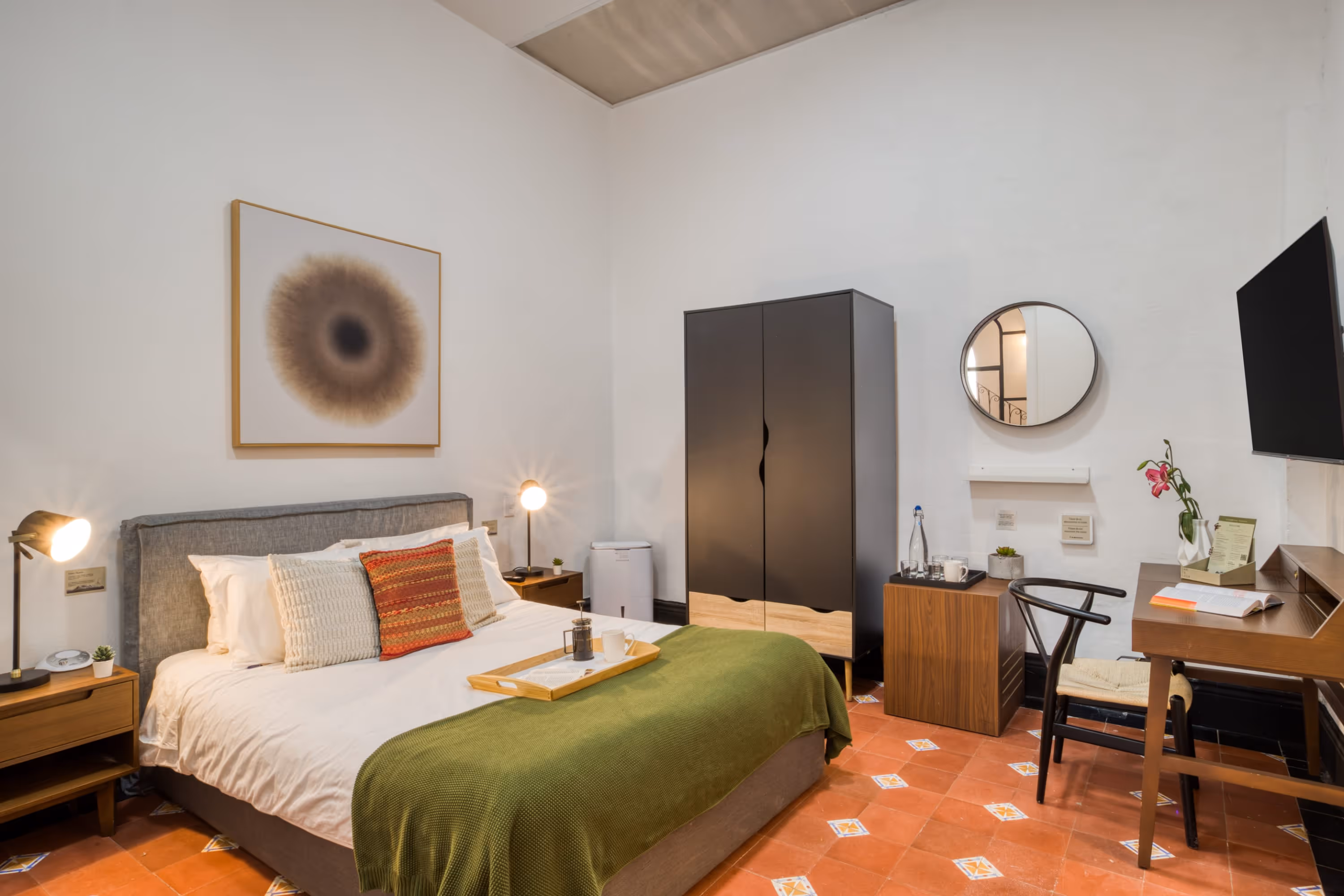 Modern bedroom with a queen-sized bed with green blanket, bedside tables with lamps, black wardrobe, round wall mirror, wooden desk with chair, and a mounted TV.