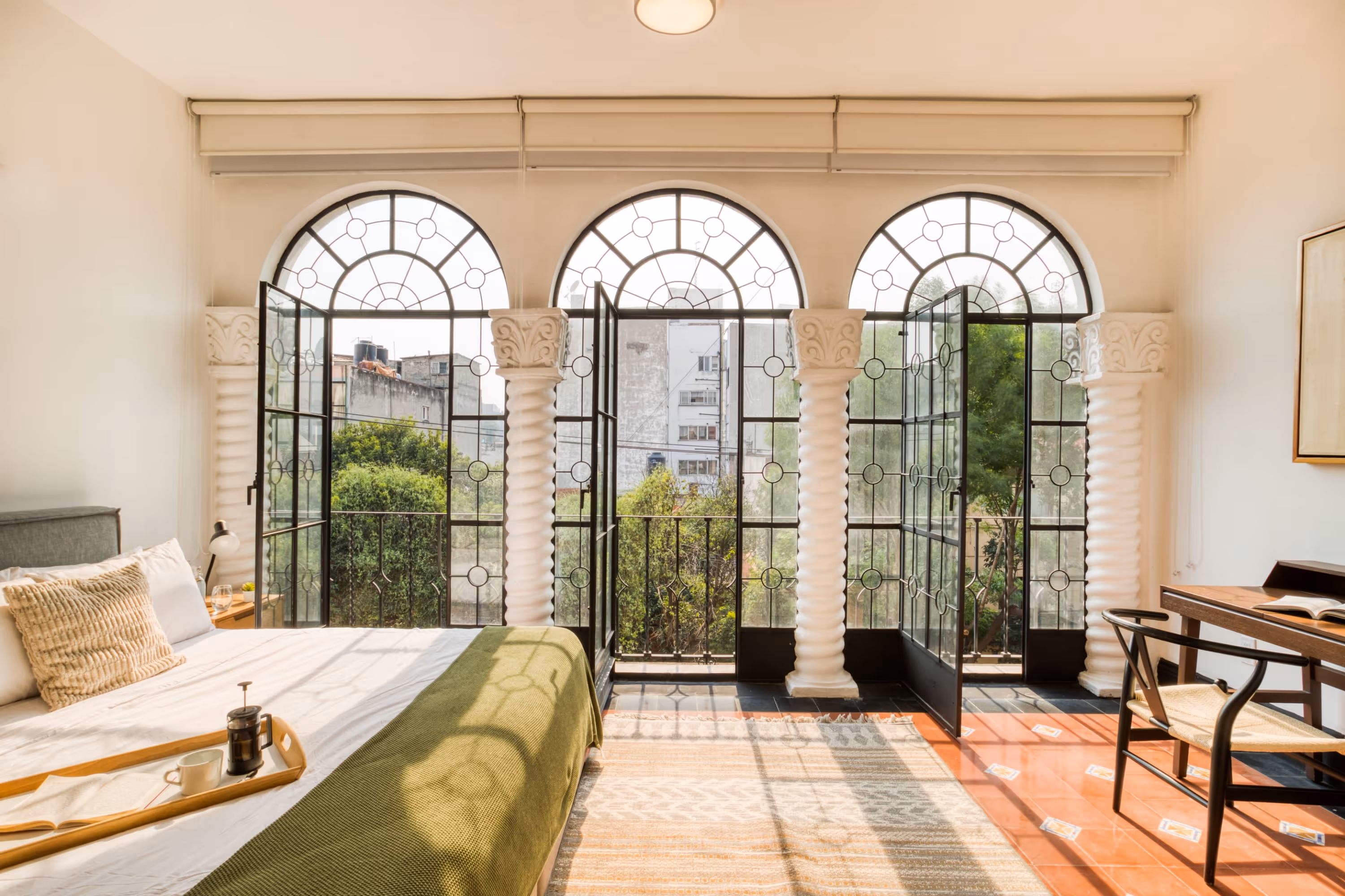 Bright bedroom with large arched windows, ornate white columns, a bed with green throw, and a wooden desk with chair.