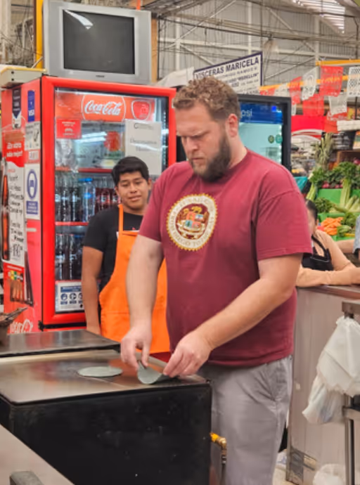 Man in a maroon shirt preparing tortillas on a griddle inside a market, with a man in an orange apron standing nearby.