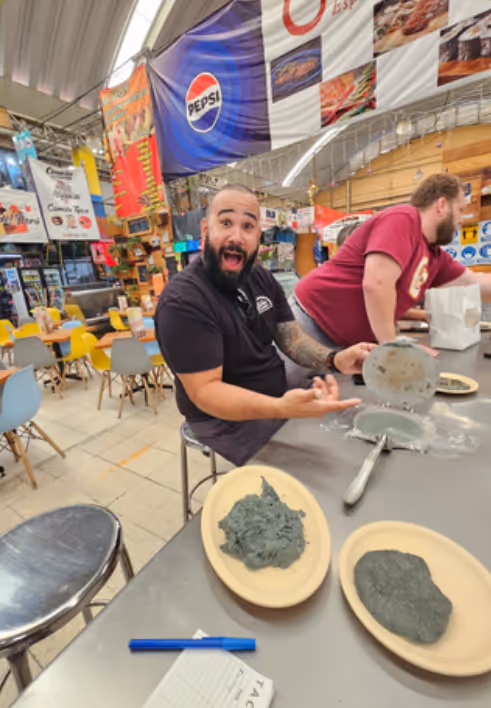 Man with a surprised expression sitting at a counter with two plates of gray dough-like substance in a casual dining area decorated with colorful banners.