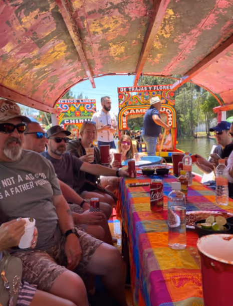 Group of people enjoying a boat ride on a colorful trajinera with drinks on the table, under a canopy with painted floral patterns.