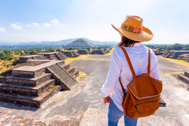 Person wearing a hat and backpack overlooking ancient pyramids at Teotihuacan under a clear blue sky.