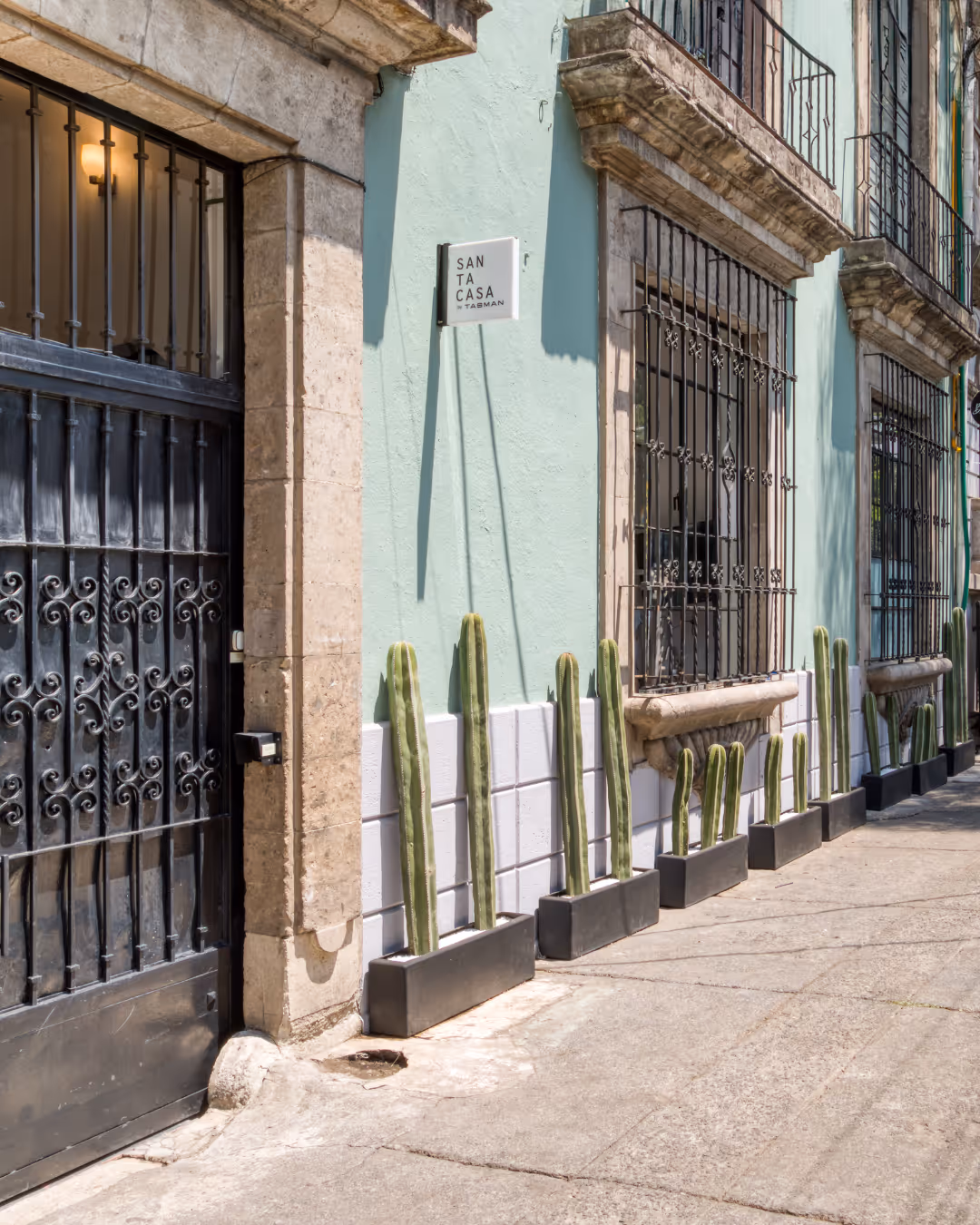 Sunlit street view of a pastel green building with black wrought iron windows, a black wrought iron gate, and tall cacti in rectangular planters along the sidewalk.