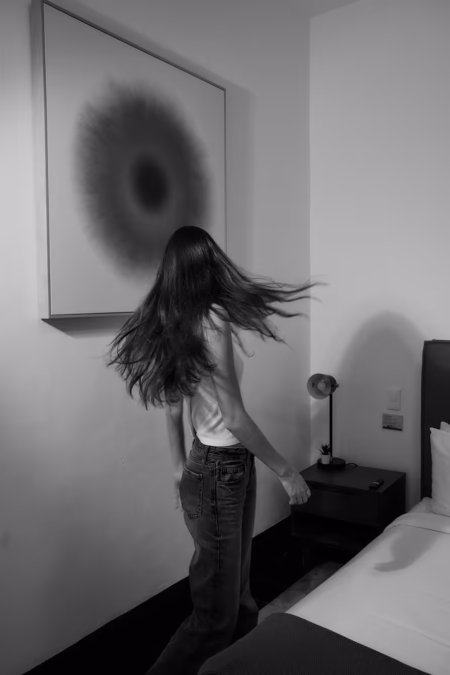 Black and white photo of a woman with long hair flipping it inside a modern bedroom near bed and nightstand.