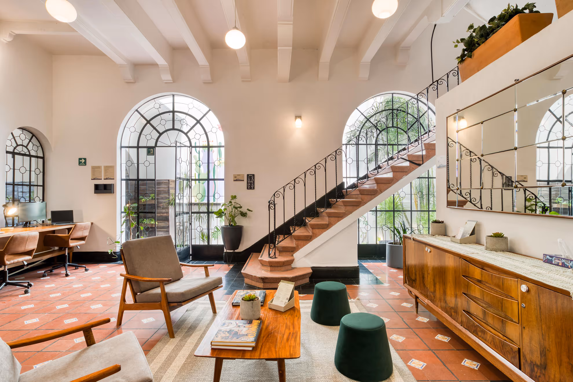 Bright living room with terracotta tiled floor, wooden furniture, green stools, large arched windows, and a staircase with wrought iron railing.
