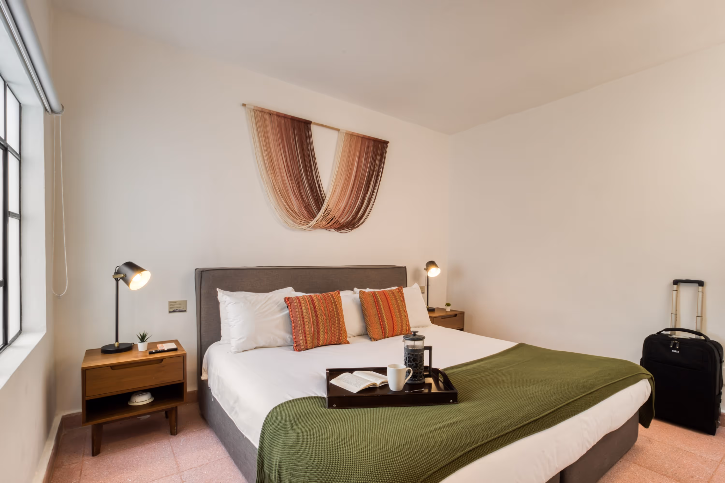Modern bedroom with a large bed featuring white linens, red patterned pillows, a green throw, wooden bedside tables with lamps, and a black suitcase near the wall.