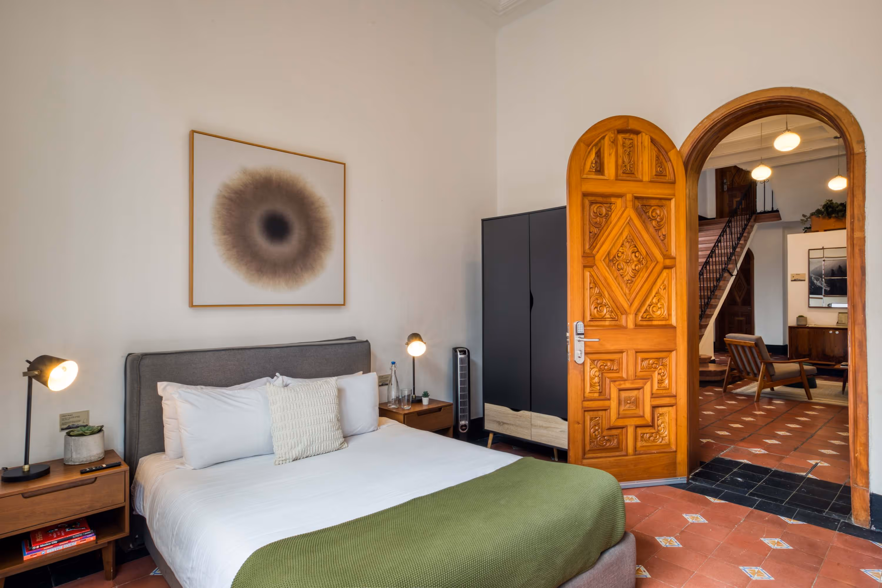 Modern bedroom with a gray upholstered bed, white and beige pillows, green blanket, wooden nightstands with lamps, and an ornate wooden door opening to a living area.