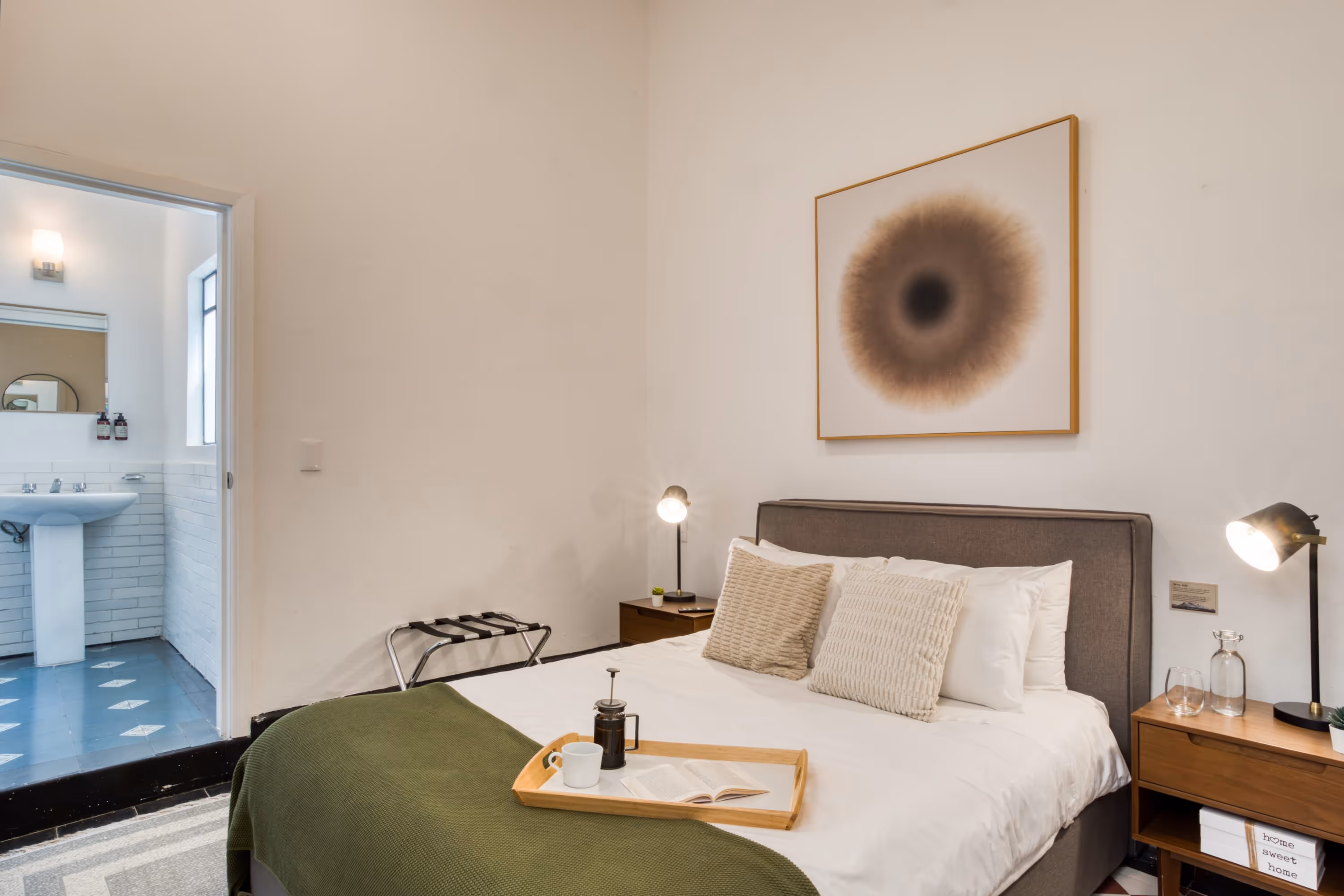 Modern bedroom with a gray upholstered bed, textured pillows, green throw blanket, wooden nightstand, and a doorway leading to a bathroom.