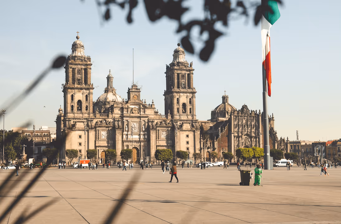 Historic Mexico City Metropolitan Cathedral in large open plaza with Mexican flag and people walking.