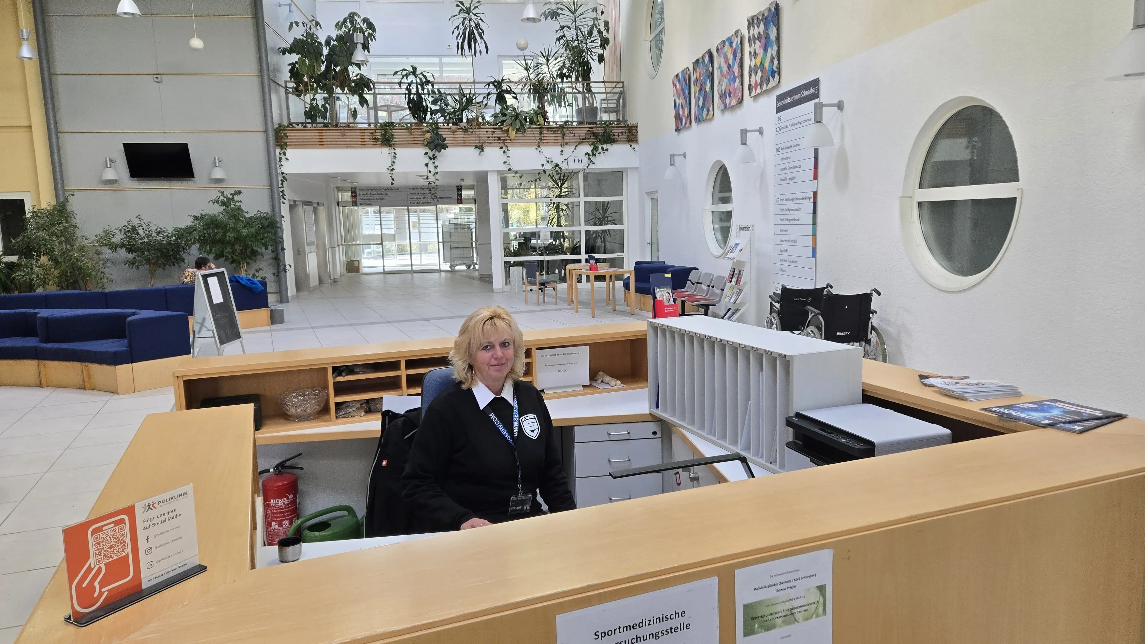 Empfangsbereich im MVZ Schneeberg: Eine Mitarbeiterin sitzt hinter einem hellen Empfangstresen und blickt in Richtung Kamera. Der offene, helle Innenraum zeigt Sitzgelegenheiten, Pflanzen, Wegweiser und Informationsmaterial. Der Bereich wirkt ruhig, übersichtlich und barrierearm gestaltet.