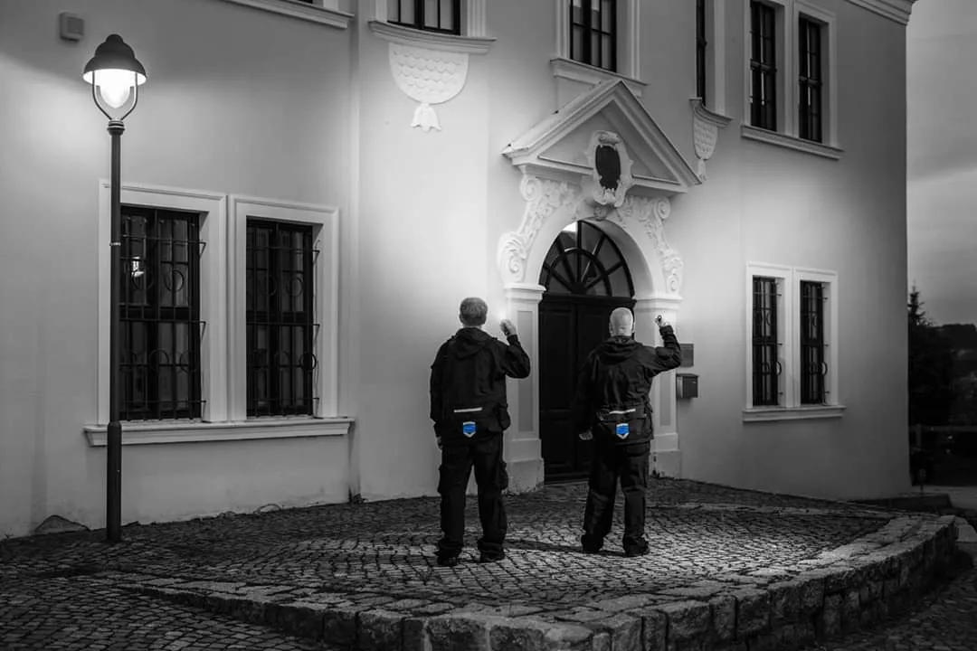 Zwei Sicherheitsmitarbeiter führen nachts einen Kontrollgang vor einem historischen Gebäude mit beleuchtetem Eingang durch.