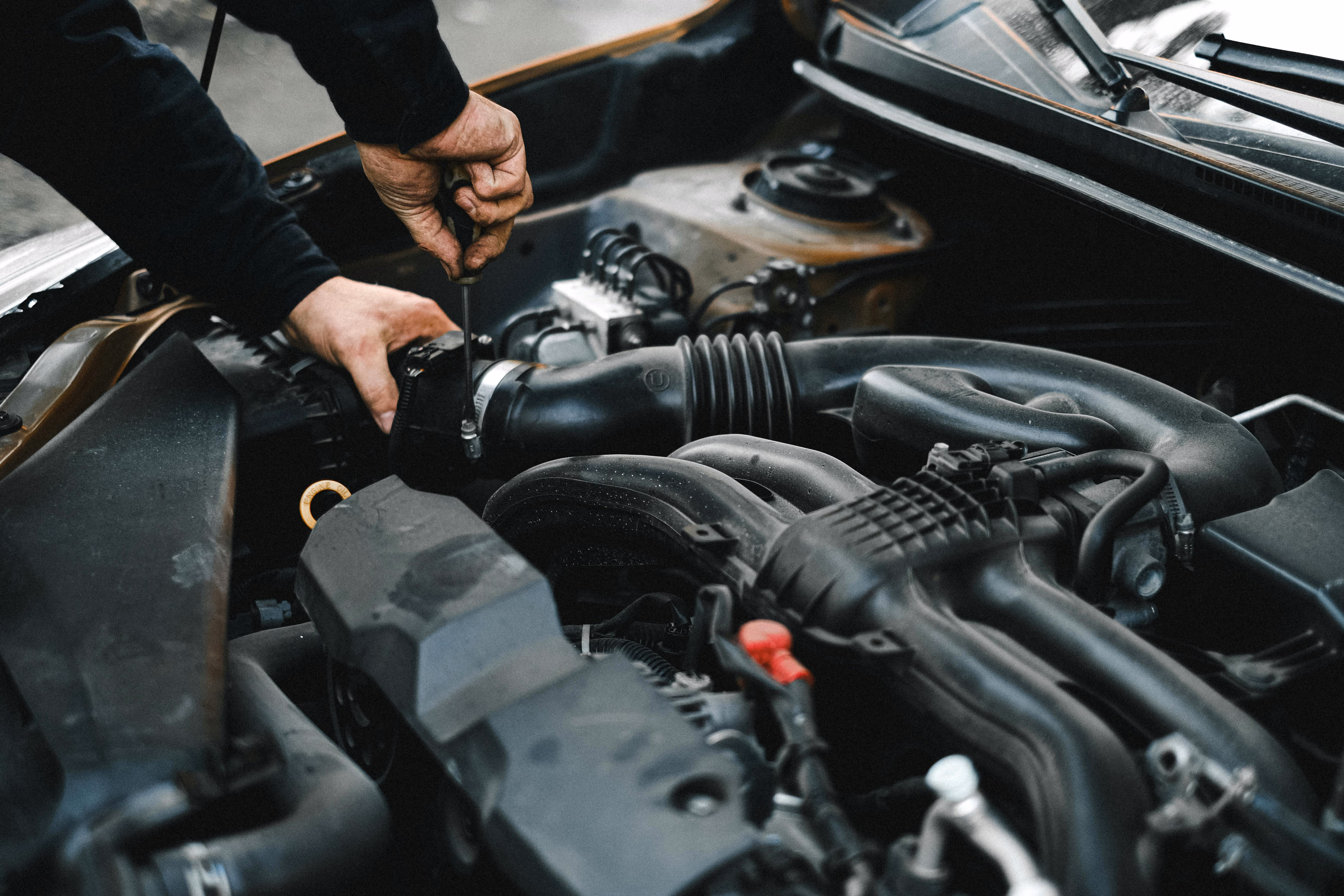 A view of underneath a car's hood showing the engine and various compartments with a mechanic's hands tightening a screw.