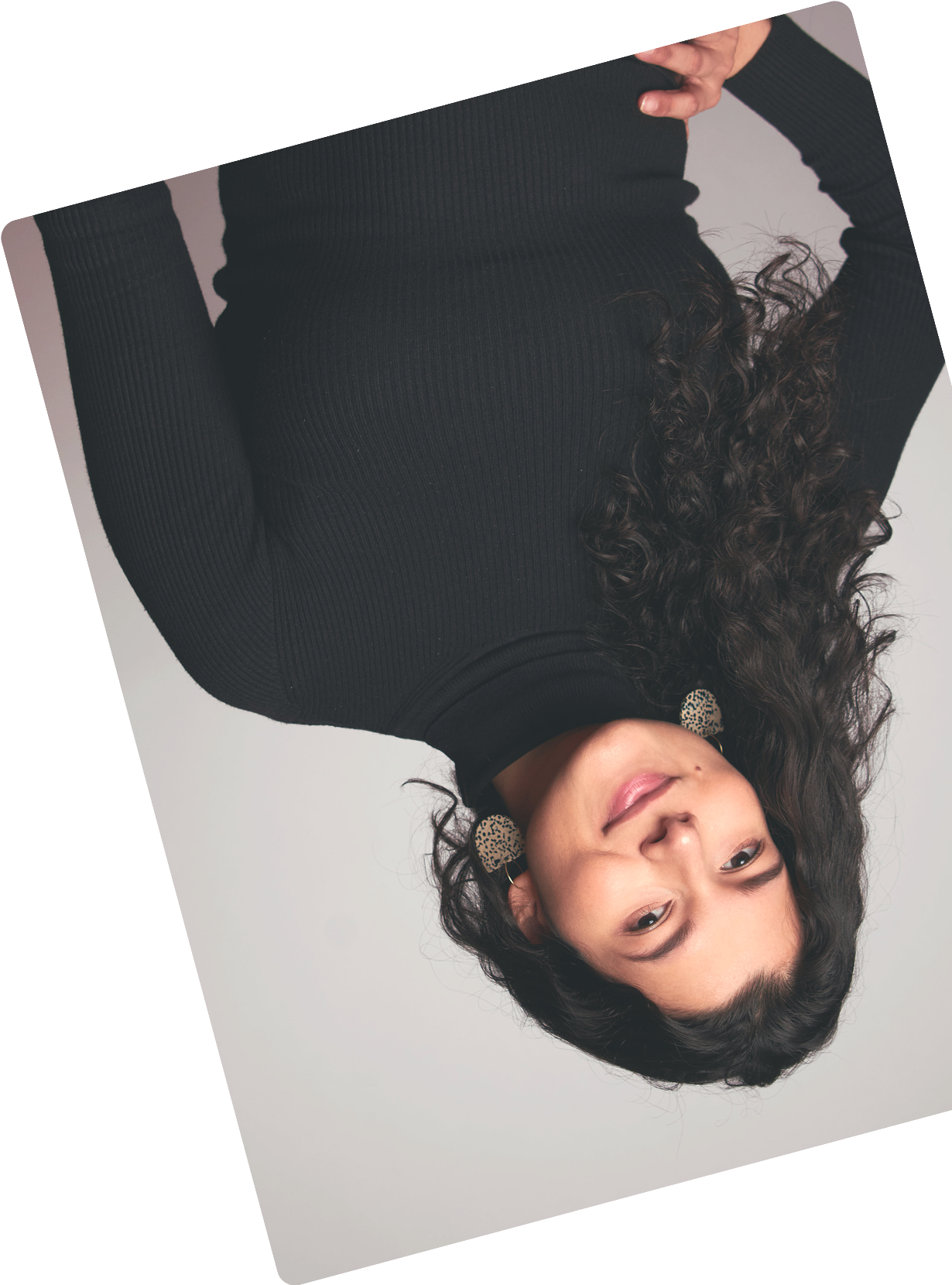 Portrait of a woman with long curly hair wearing leopard print earrings and a black ribbed turtleneck sweater against a plain background.