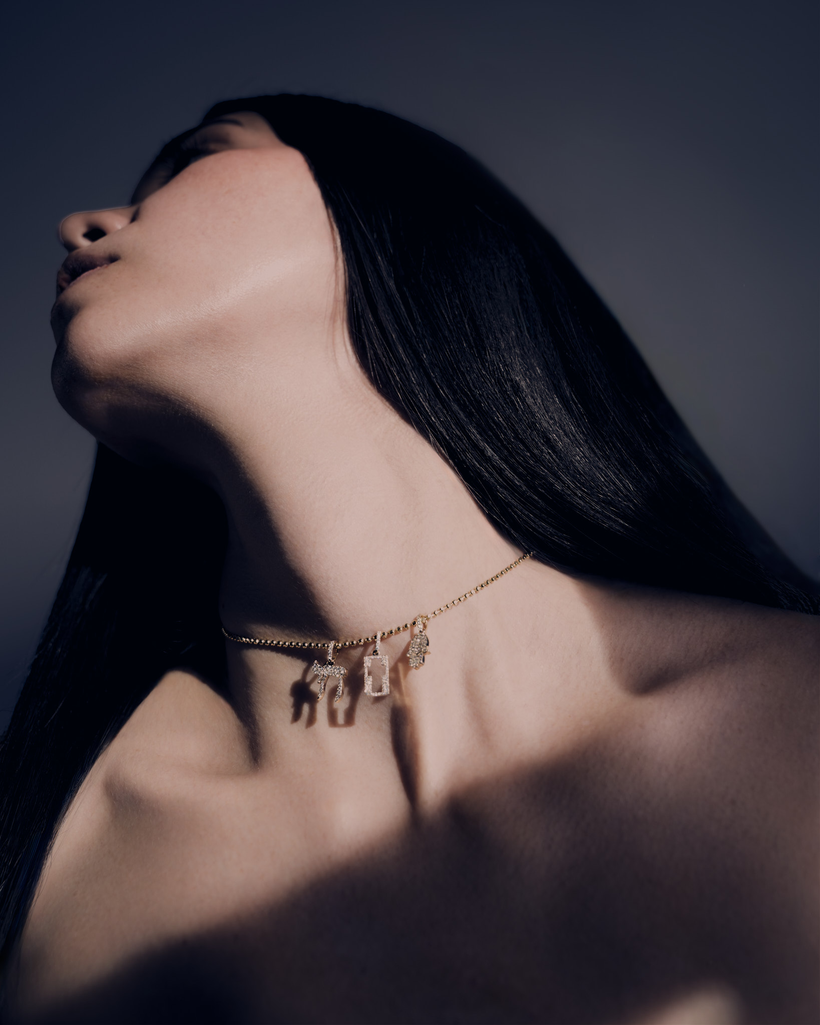 Close-up of a woman with long dark hair tilting her head back, wearing a gold necklace with four small, sparkling charms.