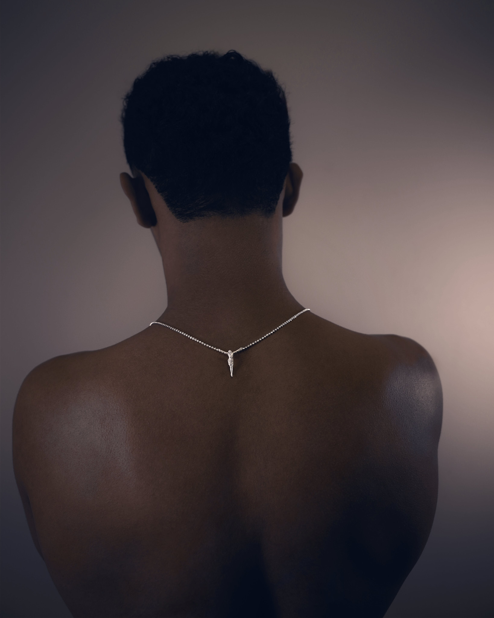 Rear view of a shirtless person with short curly hair wearing a silver necklace with a small crucifix pendant.