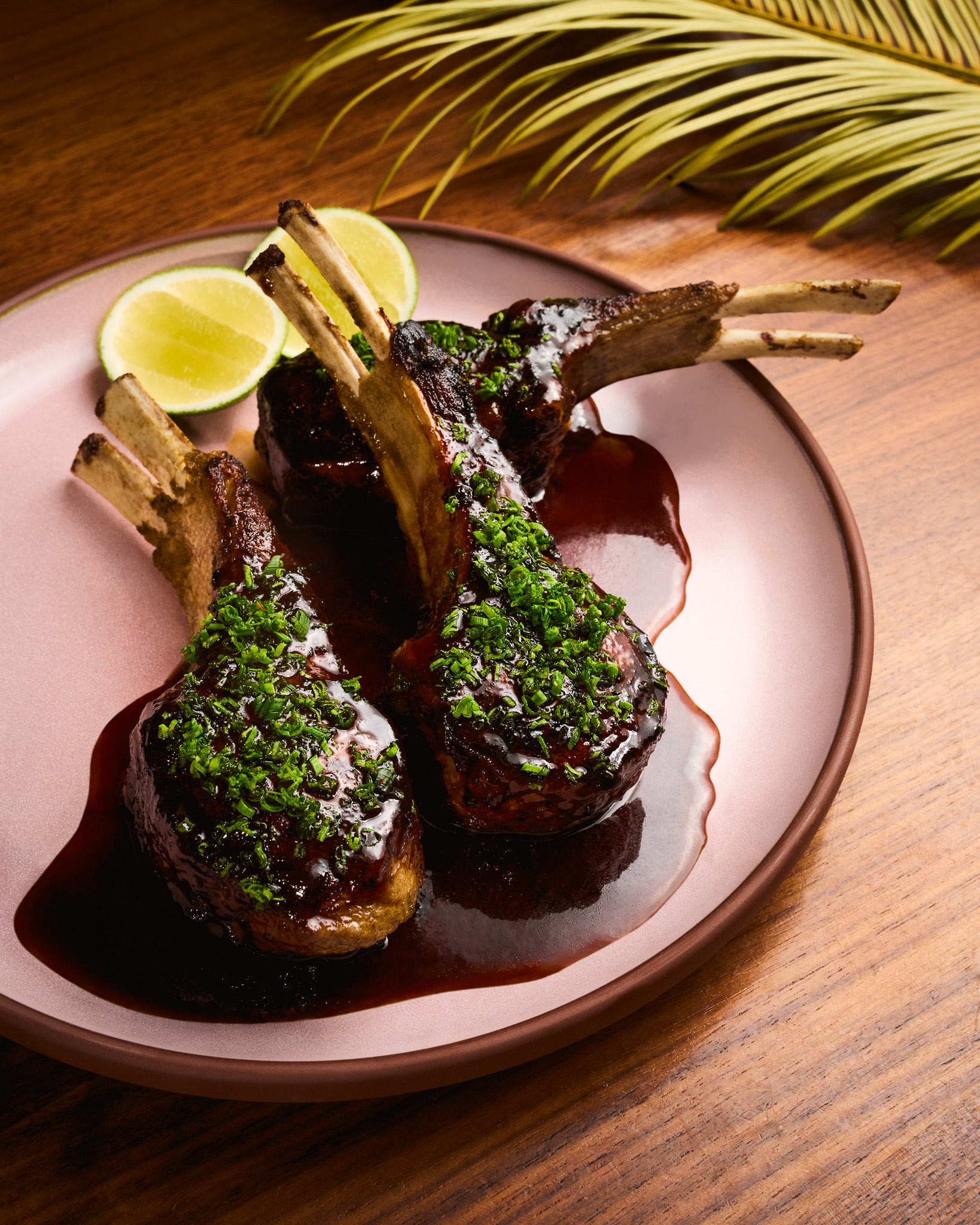 Three grilled lamb chops garnished with chopped herbs and served with dark sauce and lime wedges on a pink plate.