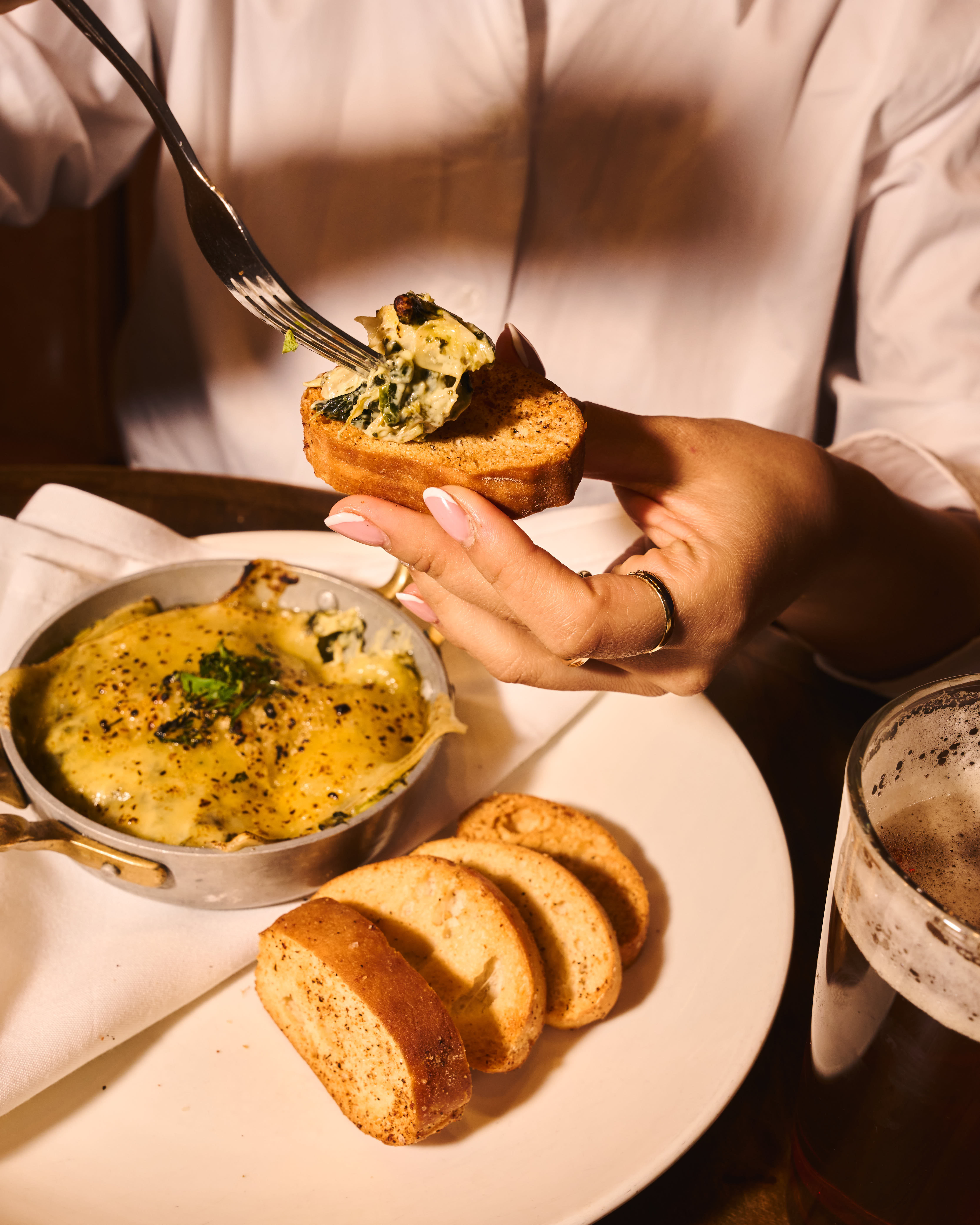 Person spreading cheesy spinach dip on toasted bread with a fork, with additional toasted bread slices and a glass of beer on the table.