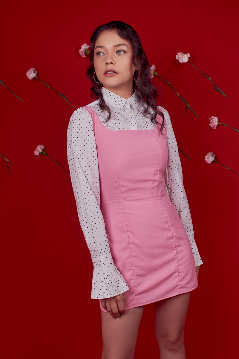 Young woman with curly hair wearing a white polka-dot blouse and pink dress, standing against a red background with pink flowers.