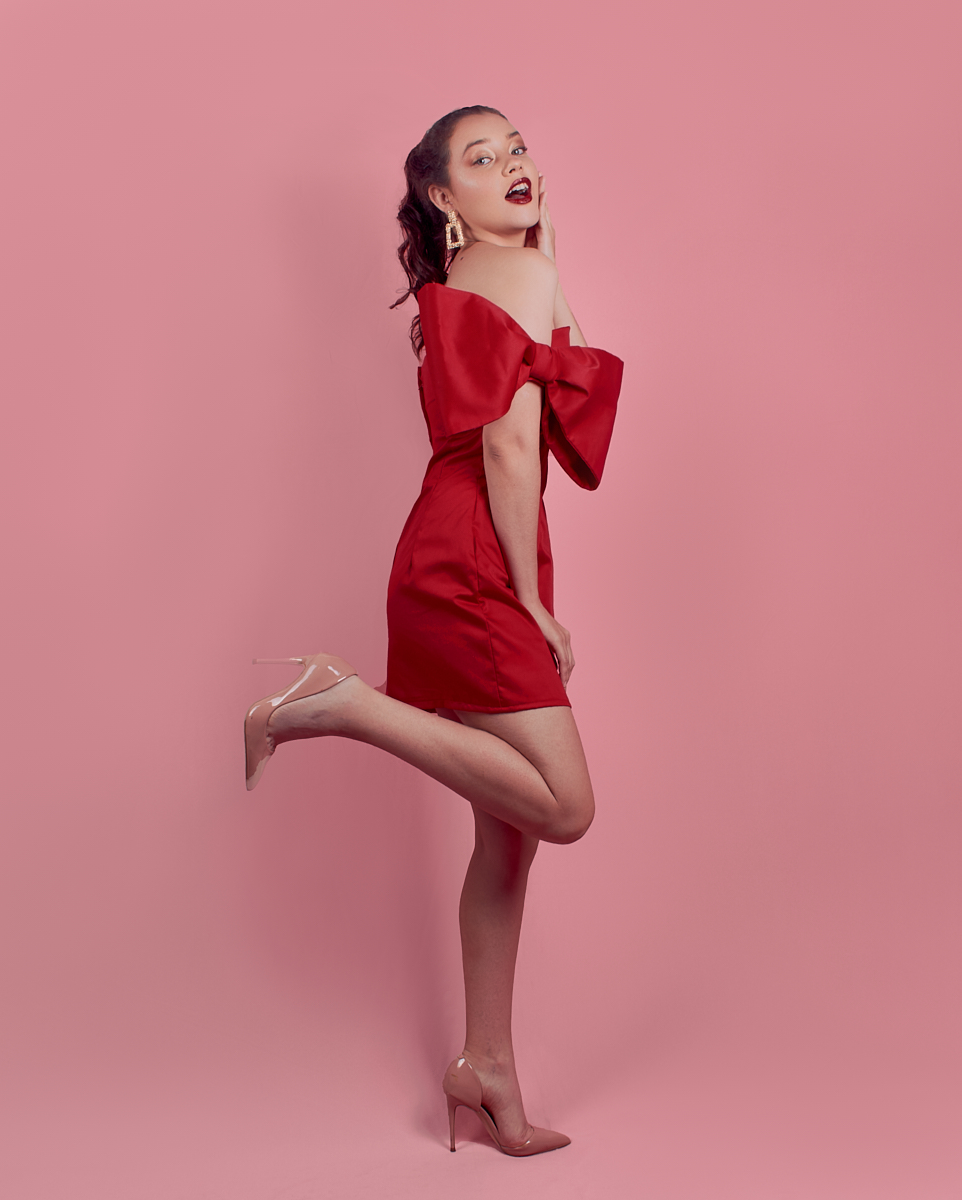 Young woman in a red off-shoulder dress with a large bow pose playfully on one leg against a pink background.