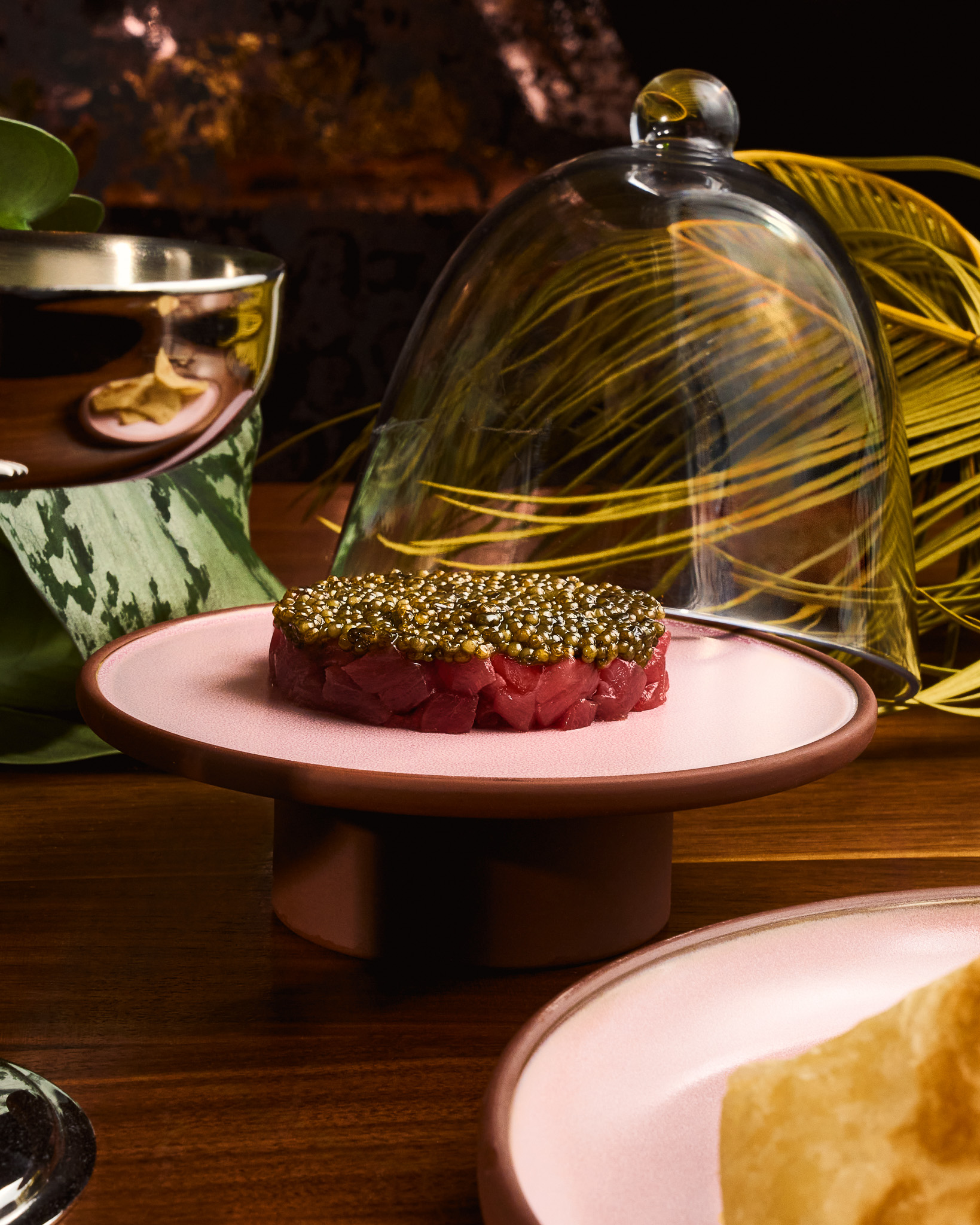 Round portion of finely diced raw tuna topped with caviar on a pink ceramic pedestal plate with a glass dome lid in the background.