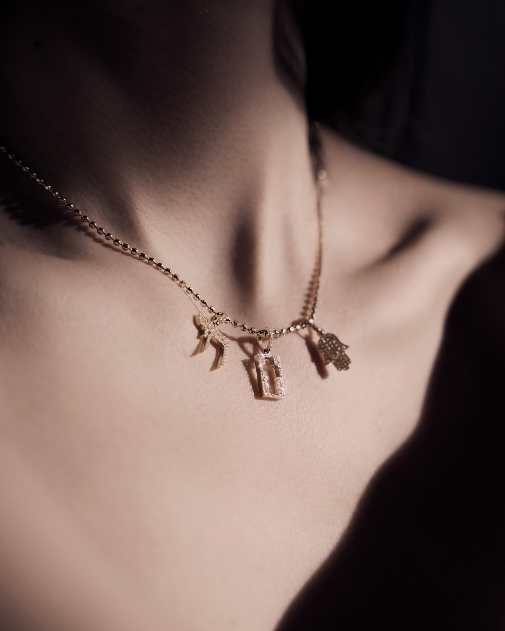Close-up of a person's neck wearing a gold necklace with three small pendants, including a Hamsa hand and a rectangular charm.