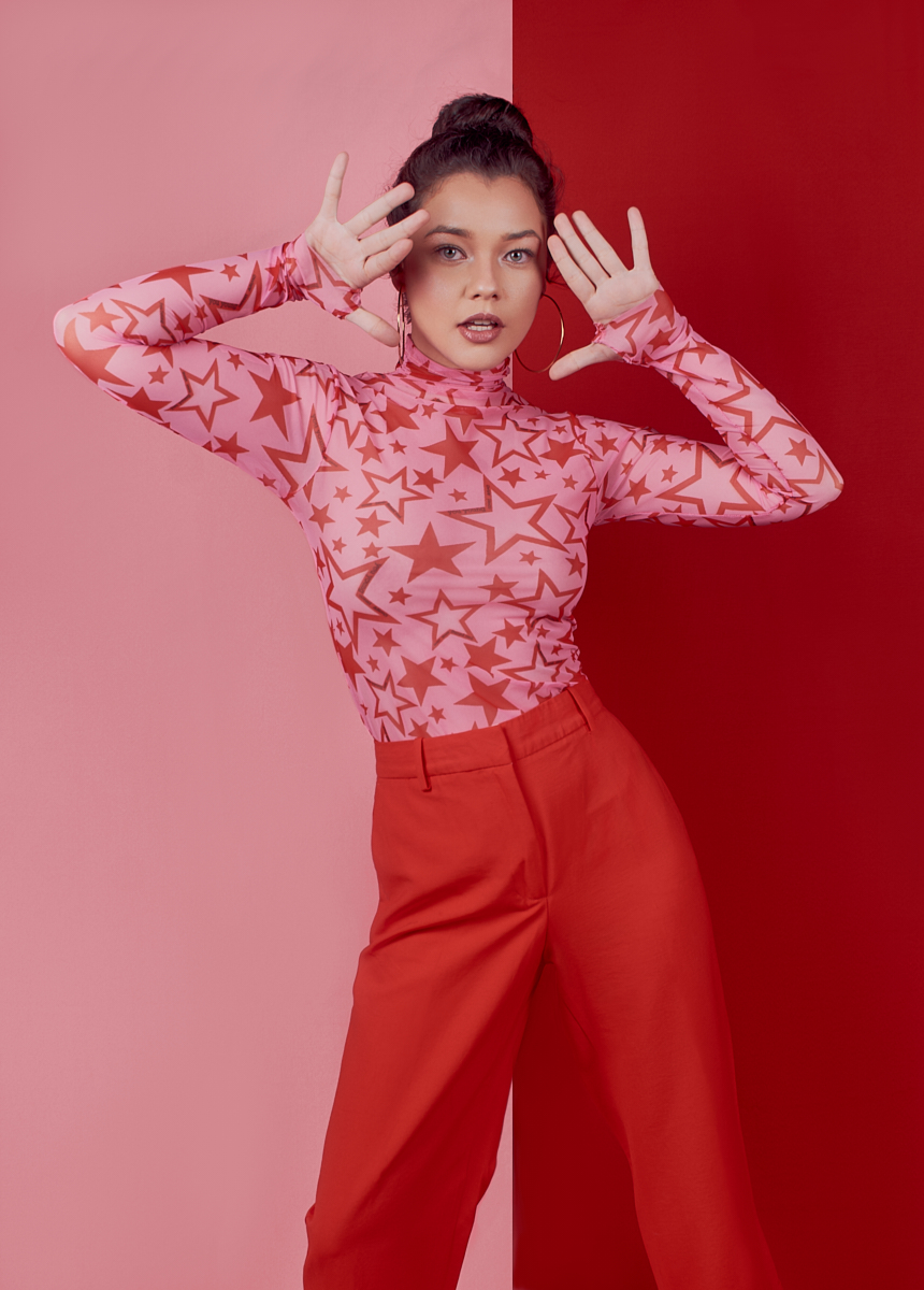 Woman wearing a pink top with red stars and red pants posing with hands near her face against a half pink, half red background.