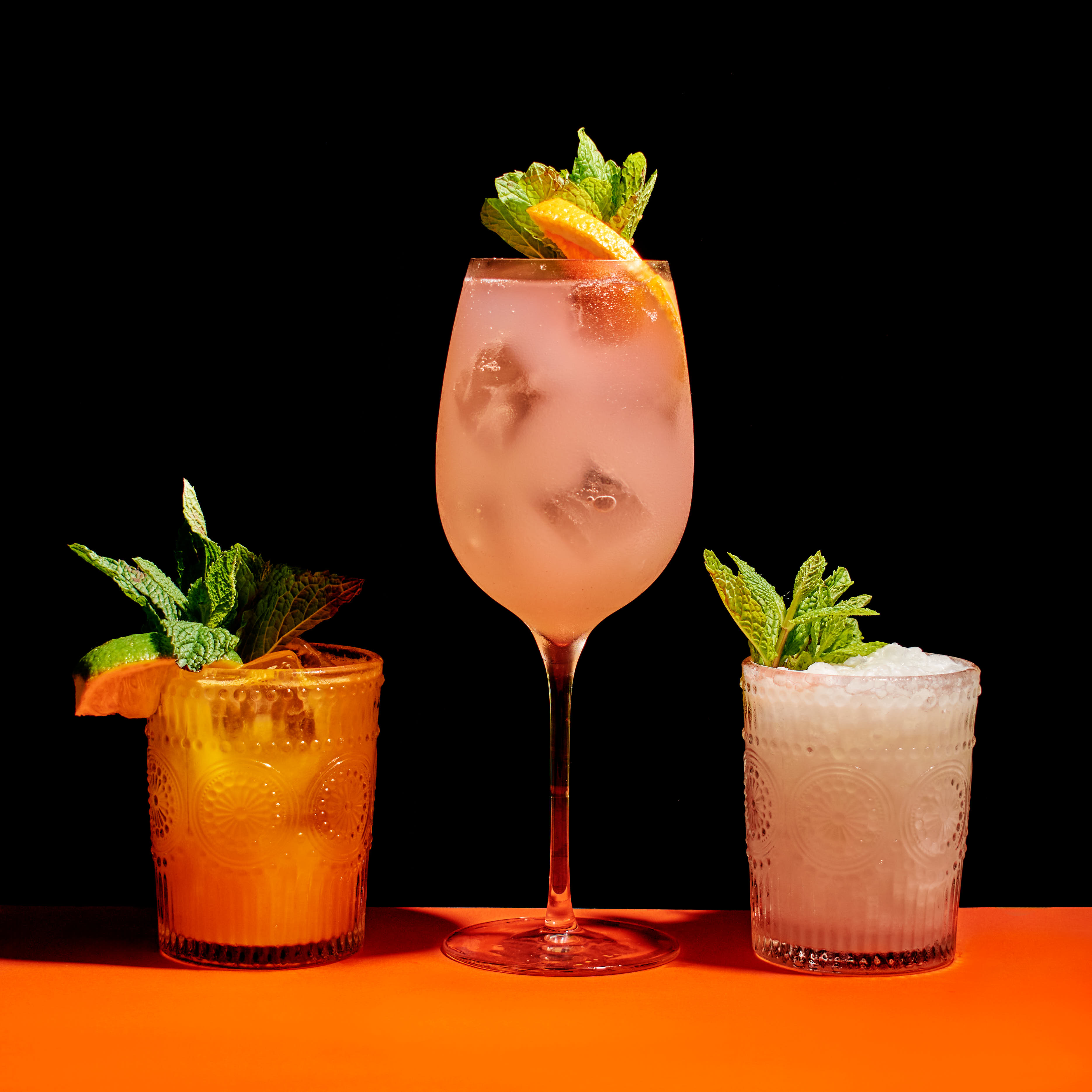 Three colorful cocktails with mint garnish, ice, and citrus slices on an orange surface against a black background.