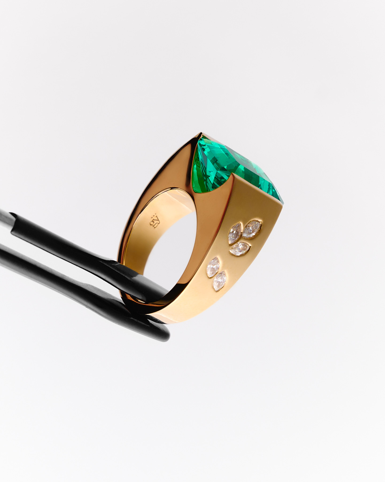 Gold ring with a large square green gemstone and six small marquise-cut diamonds held by tweezers against a white background.