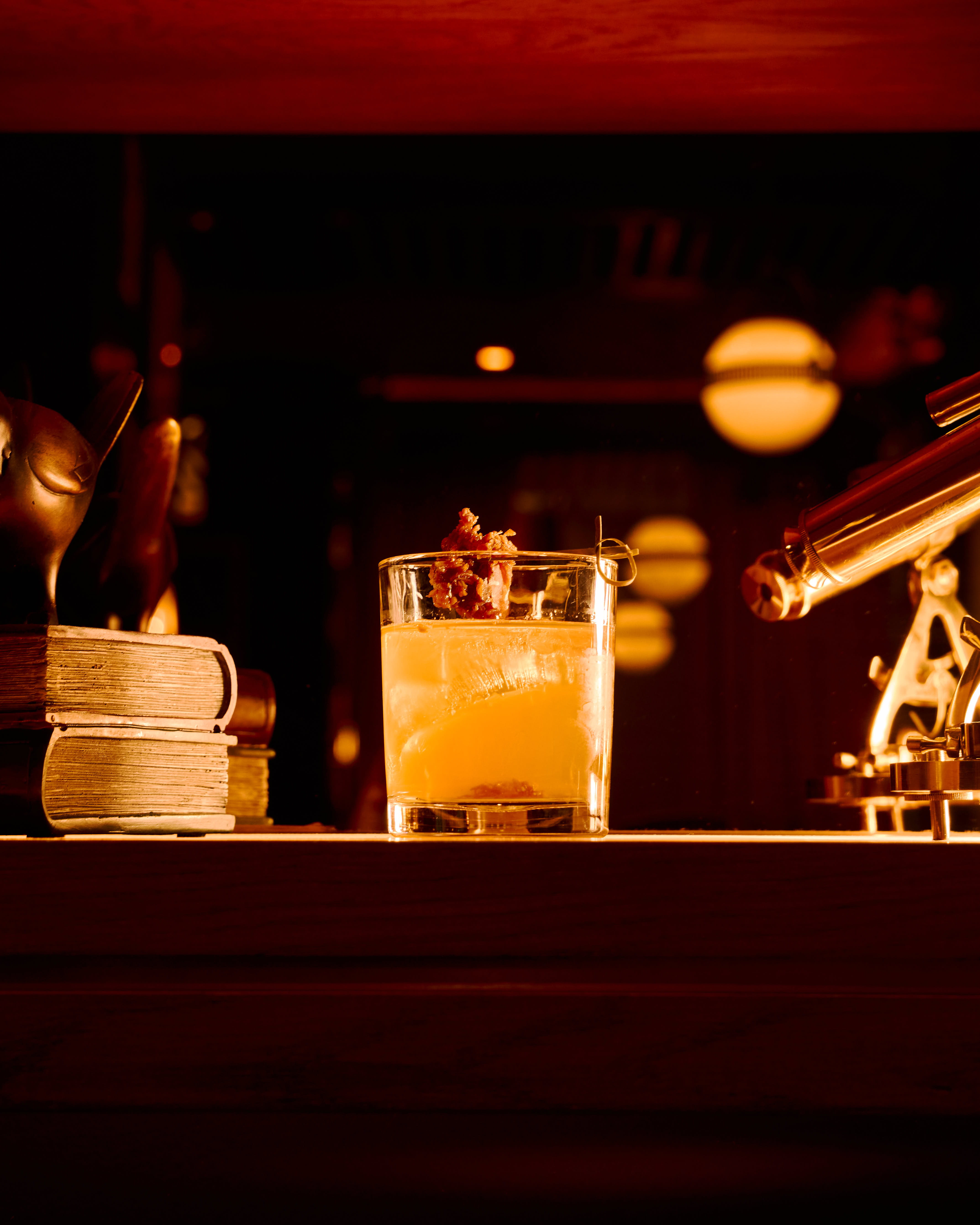 Glass of amber cocktail with ice and garnish on a wooden surface, flanked by old books and a brass telescope under warm lighting.
