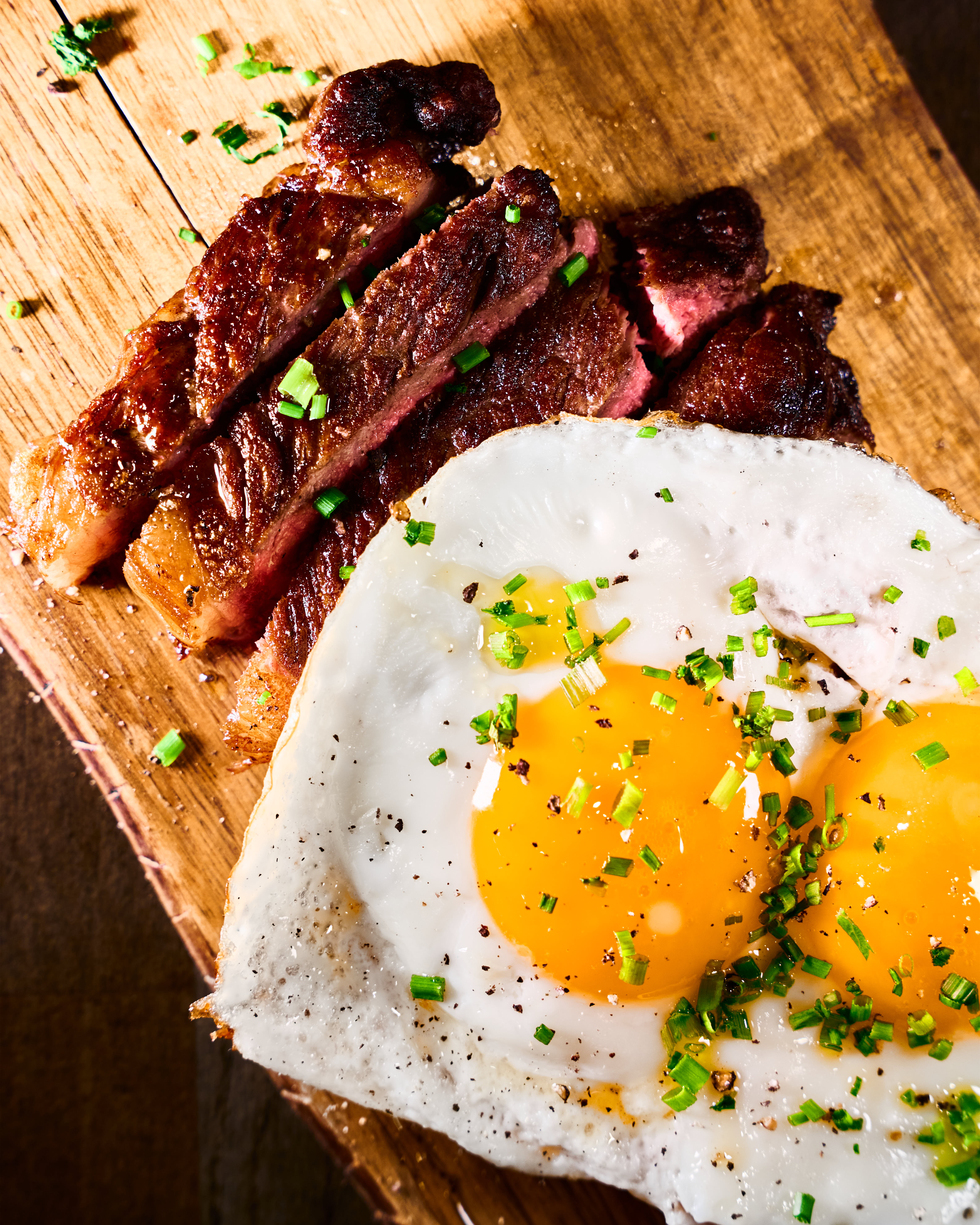 Two sunny-side-up eggs seasoned with chopped chives and black pepper next to slices of grilled steak on a wooden cutting board.