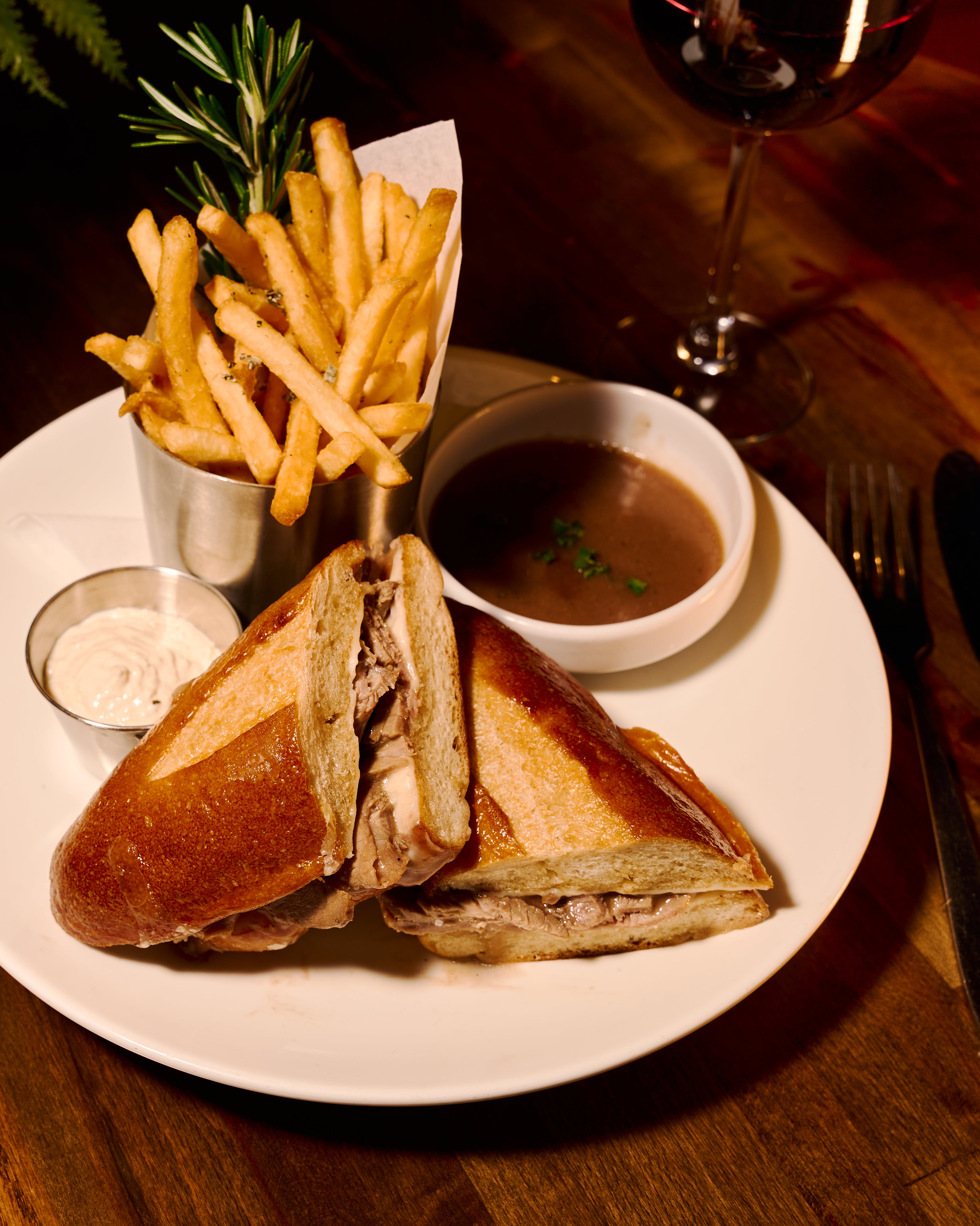 Plate with a sliced roast beef sandwich, a metal cup of French fries garnished with rosemary, a small cup of dipping sauce, and a bowl of au jus, alongside a glass of red wine on a wooden table.