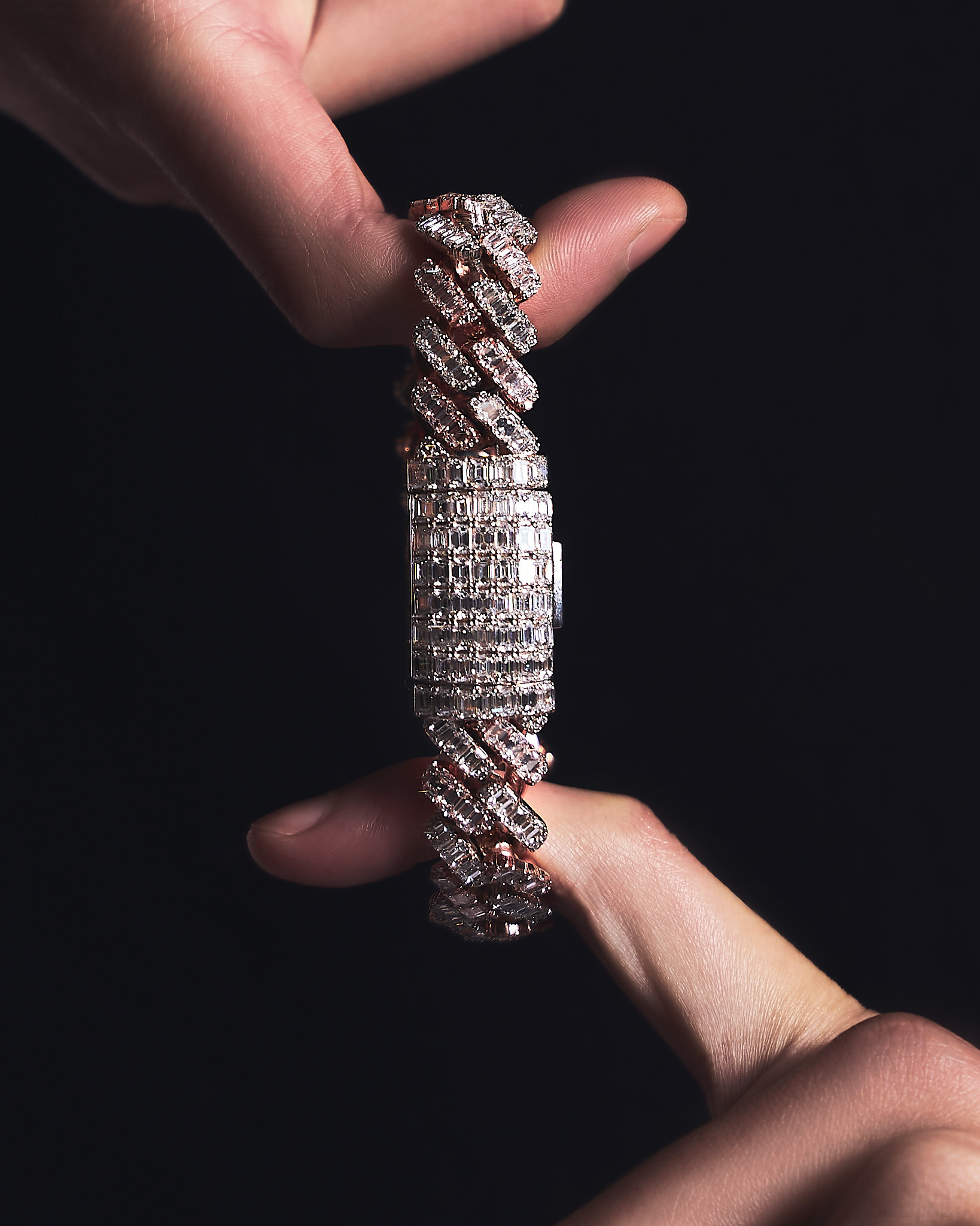 Hand holding a silver and rose gold bracelet encrusted with rectangular and square diamonds against a black background.