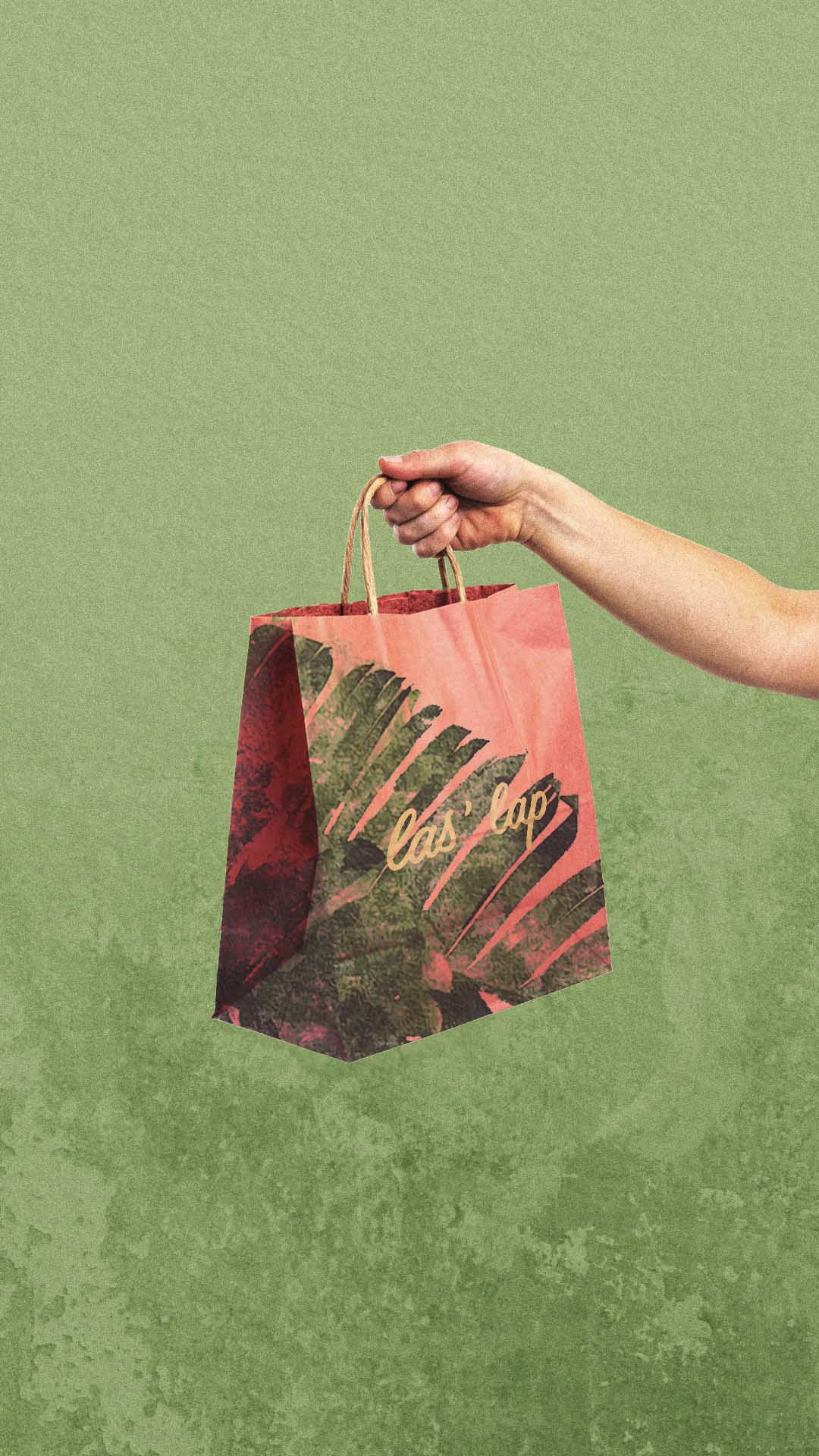Hand holding a red paper bag with green leaf patterns and the text 'las lap' on a green textured background.