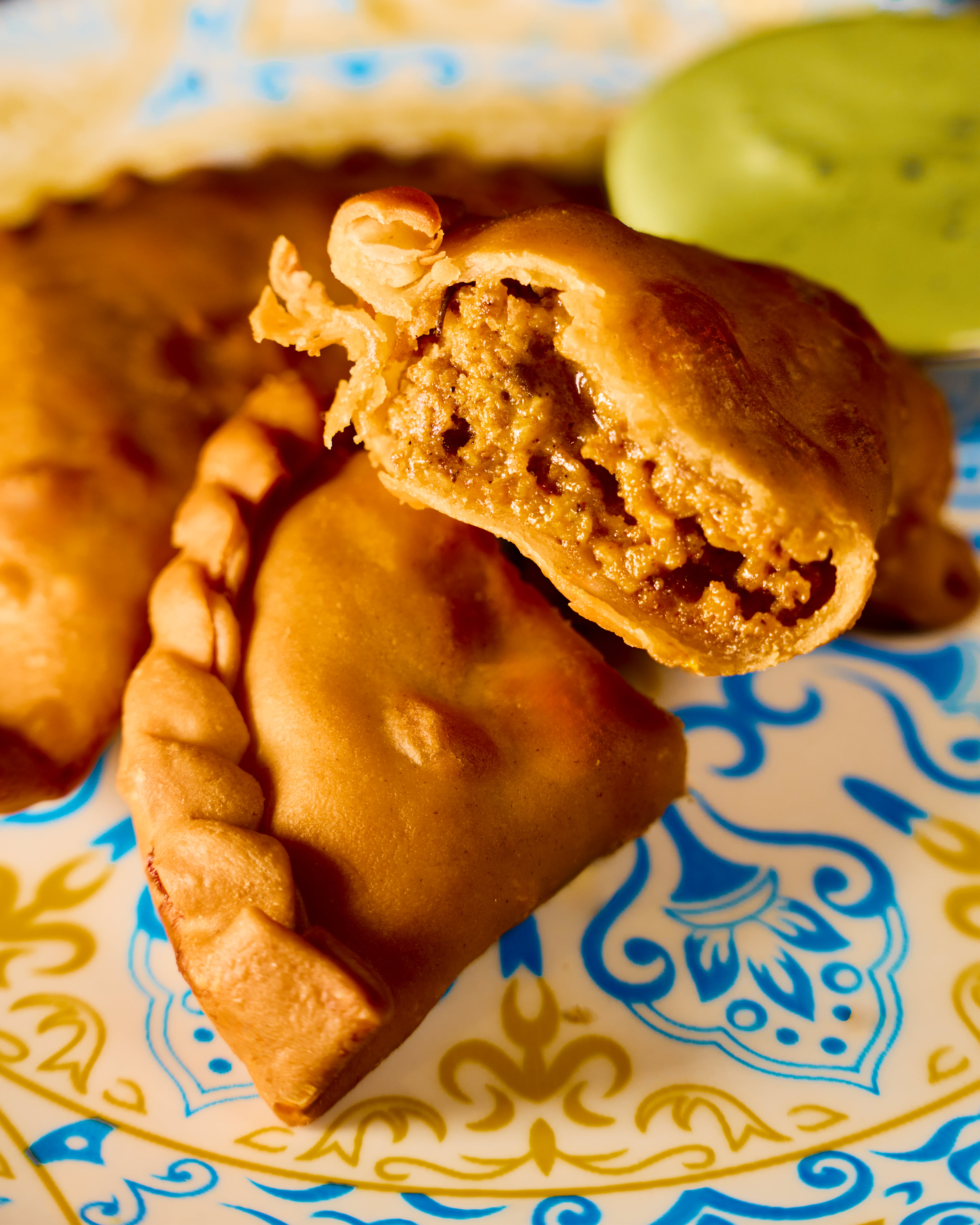 Close-up of three cooked meat-filled empanadas on a decorative blue and yellow plate with green dipping sauce in the background.