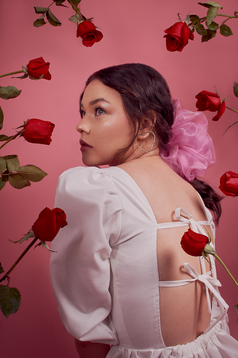 Woman with dark hair wearing a white dress with open back tied by bows, surrounded by red roses on a pink background.