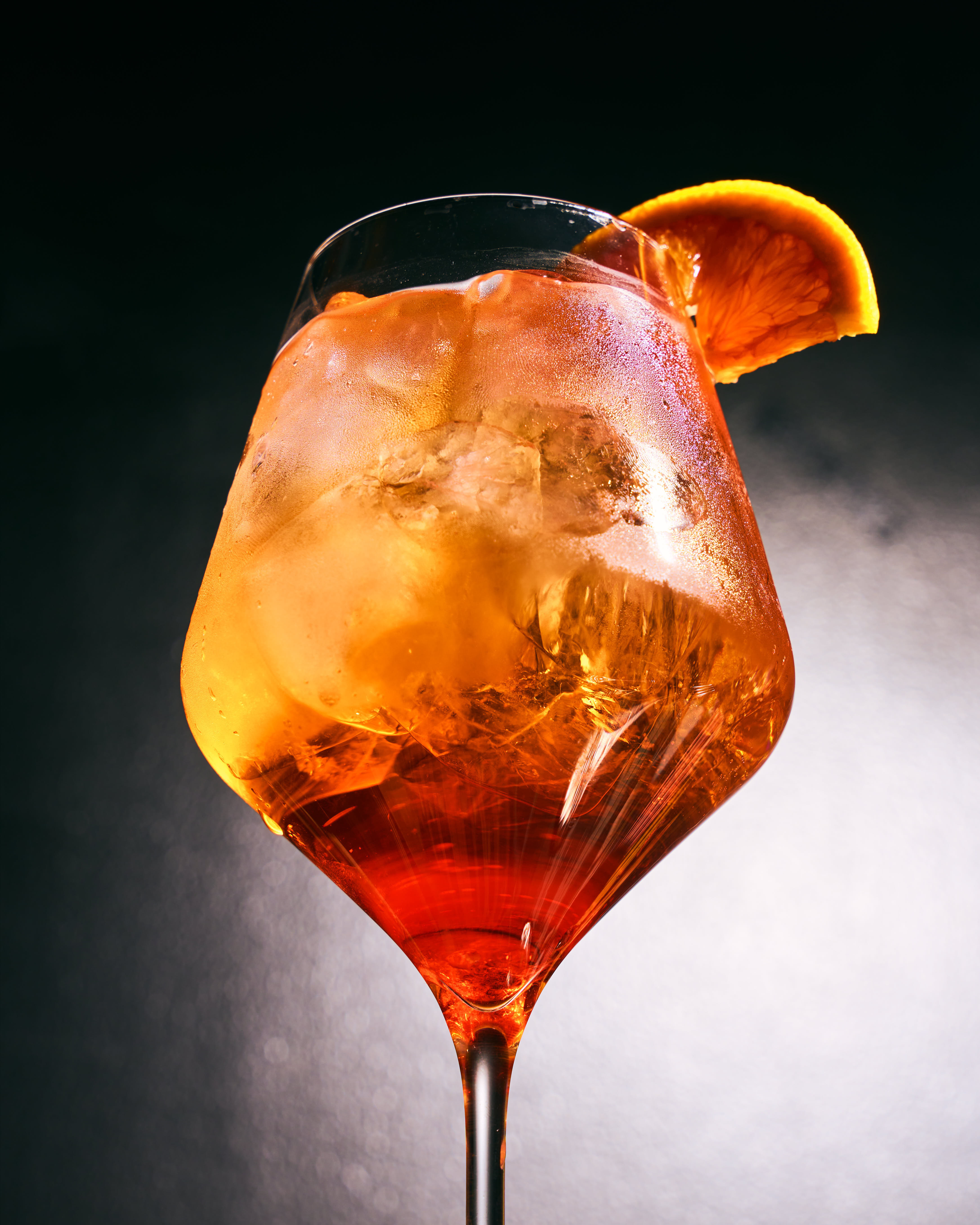 Close-up of an iced orange cocktail garnished with a slice of orange in a stemmed glass against a dark background.