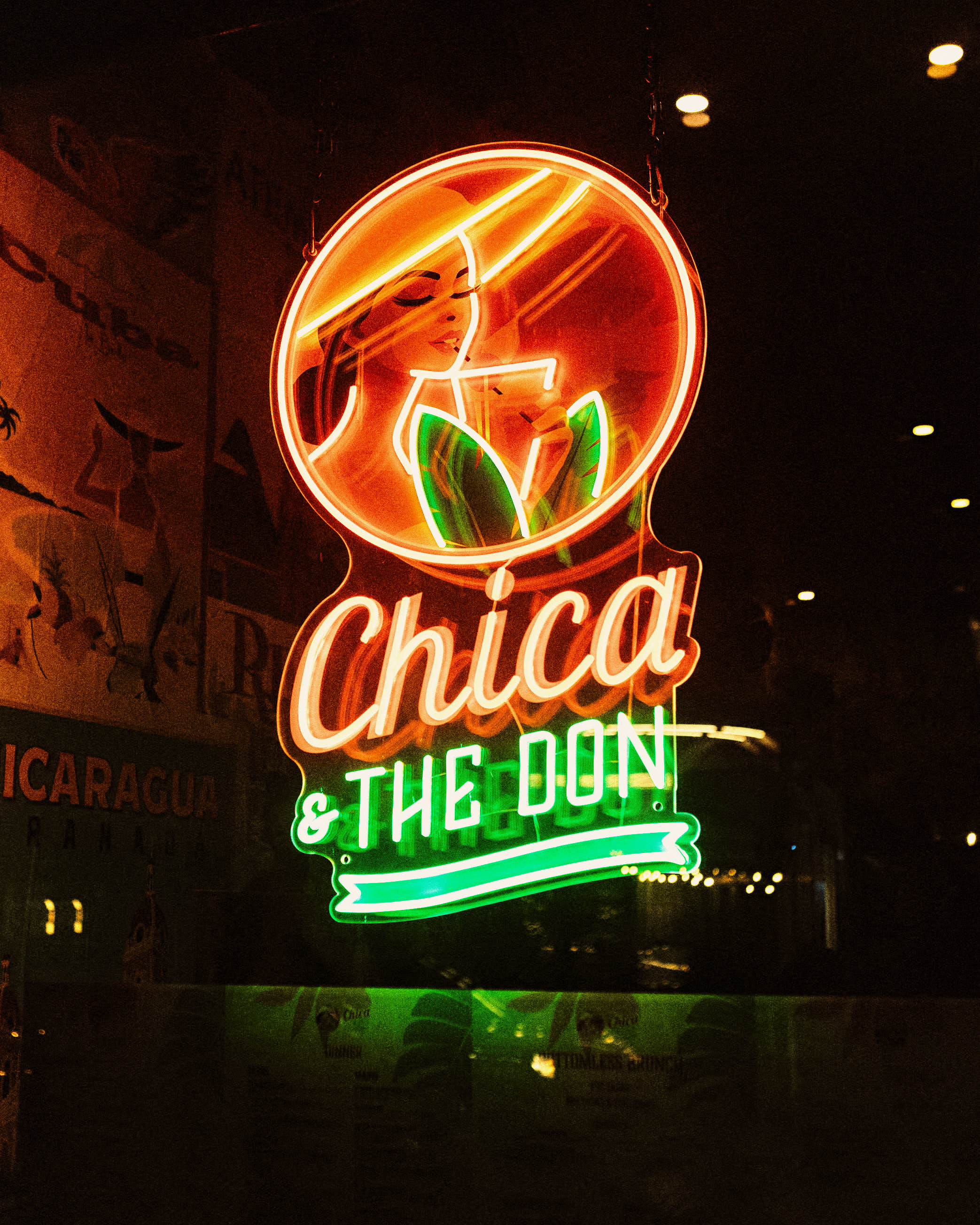 Neon sign showing a woman in a hat drinking from a glass with leaves and the text 'Chica & The Don' in bright orange and green.